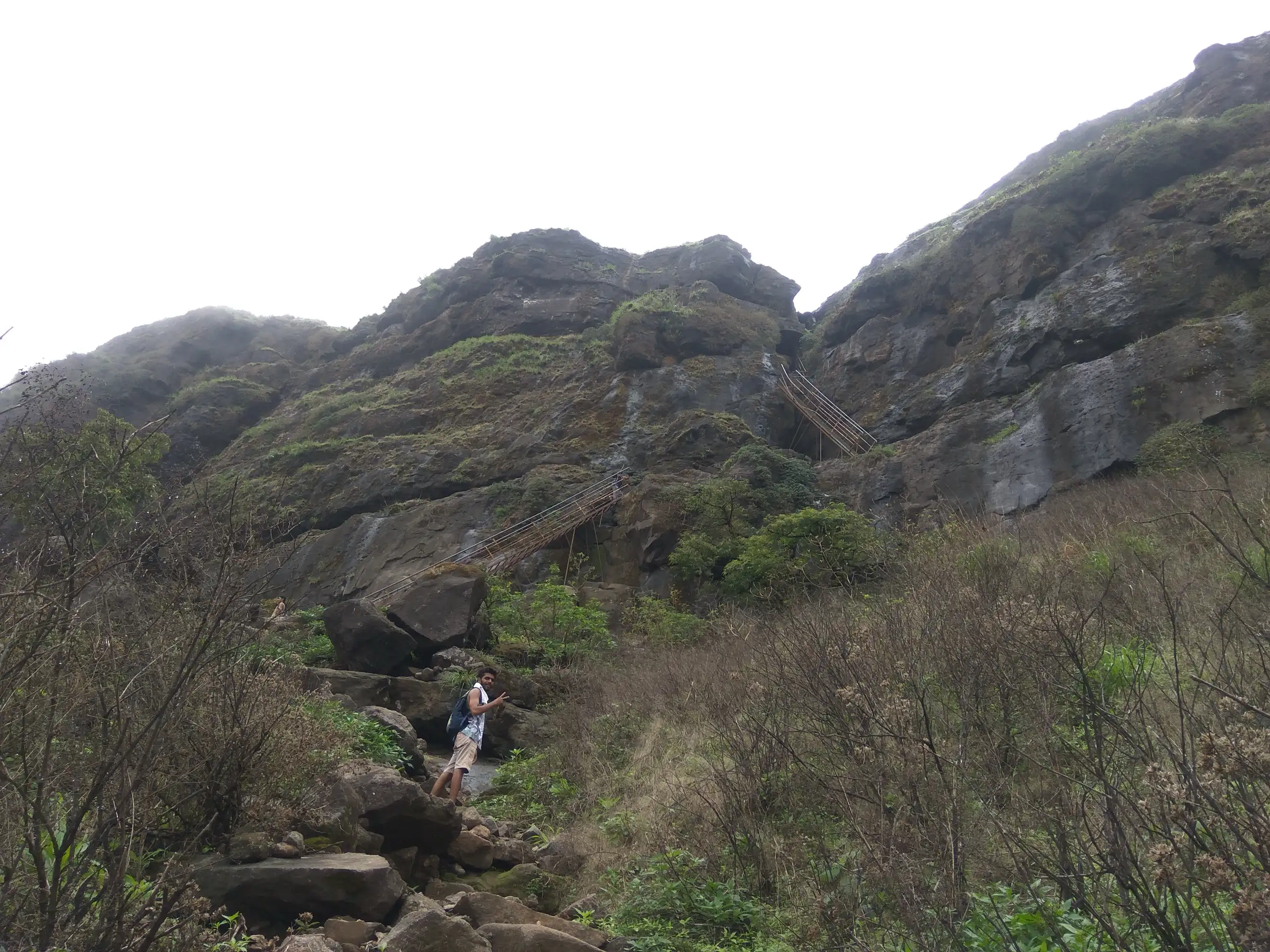 Steel ladders fixed to a mossy rock face below Ratangad, with a trekker on the boulder trail in front