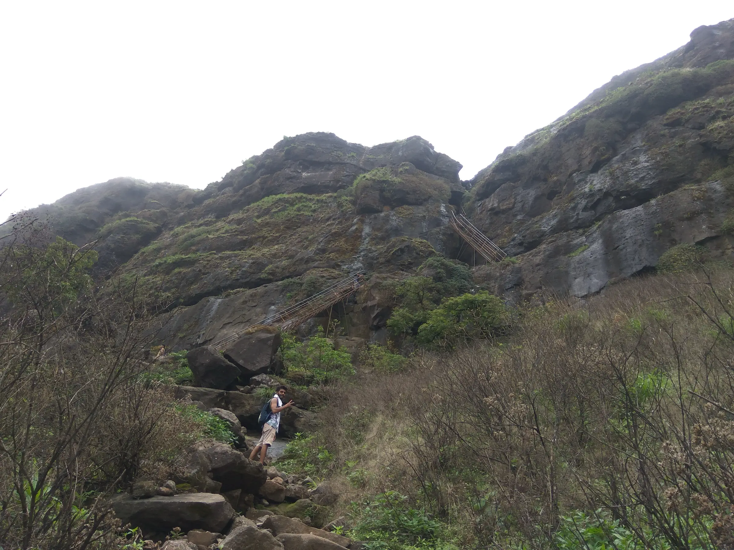 Steel ladders fixed to a mossy rock face below Ratangad, with a trekker on the boulder trail in front