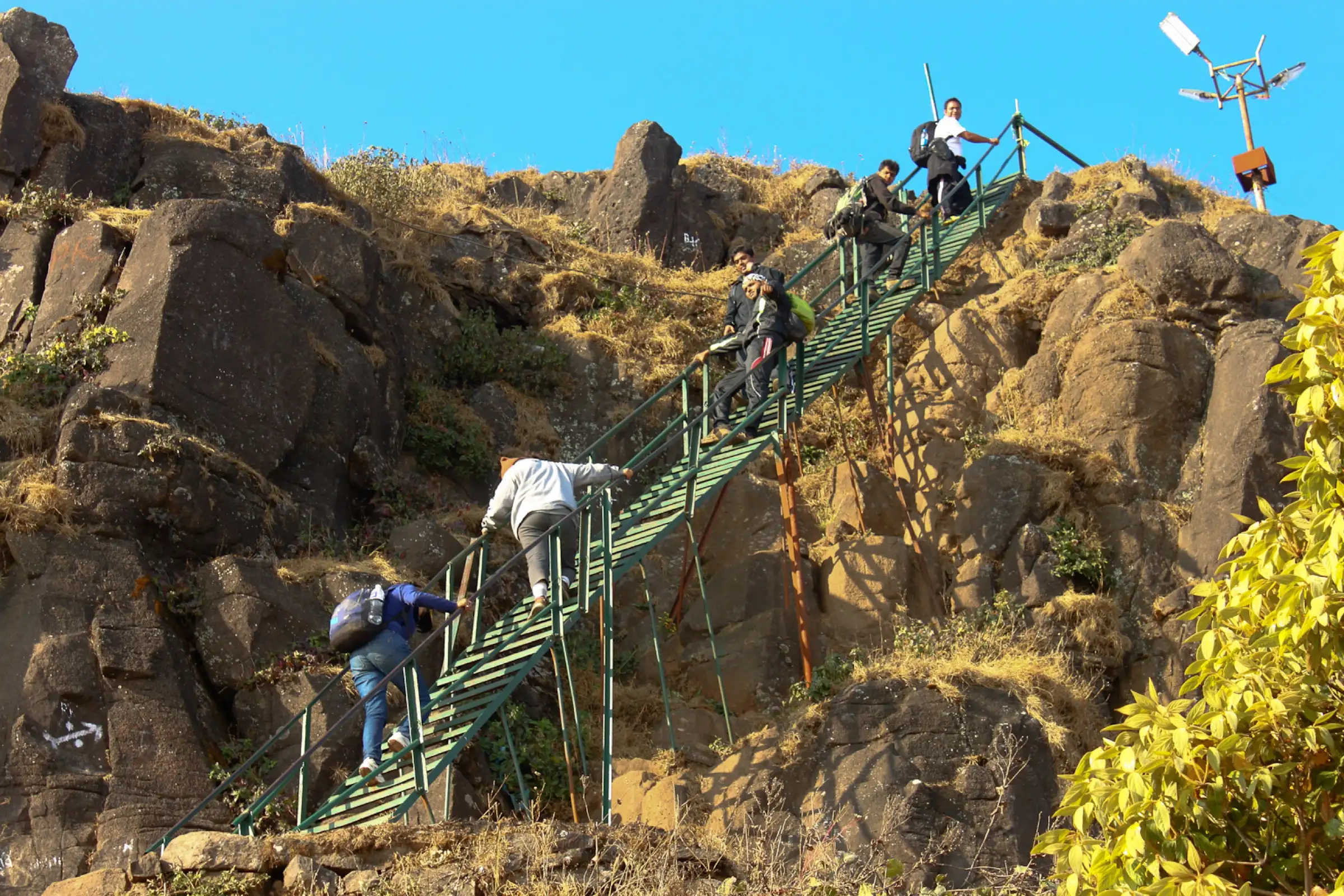 Trekkers climbing a green iron ladder up a rock band on Kalsubai