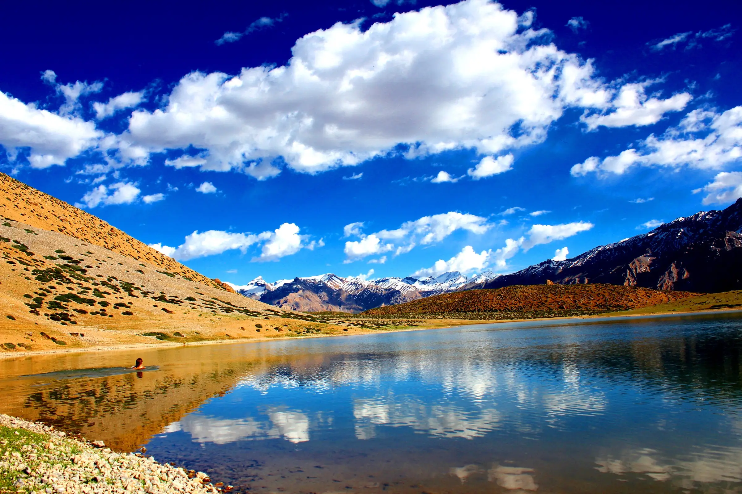 Dhankar Lake reflecting white clouds and snow peaks, with a pebbly shore and grassy brown slopes