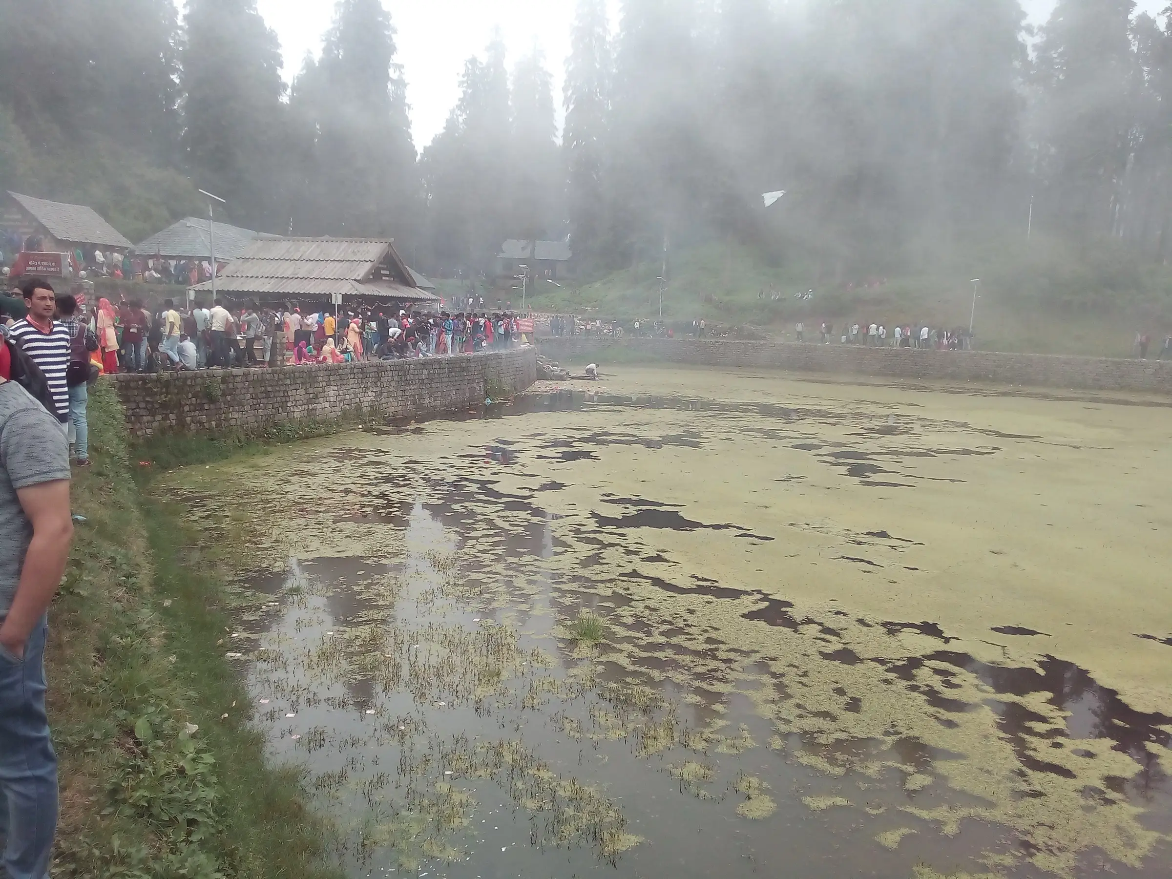 Crowds of devotees along the stone-walled edge of Kamrunag lake in mist during the June fair