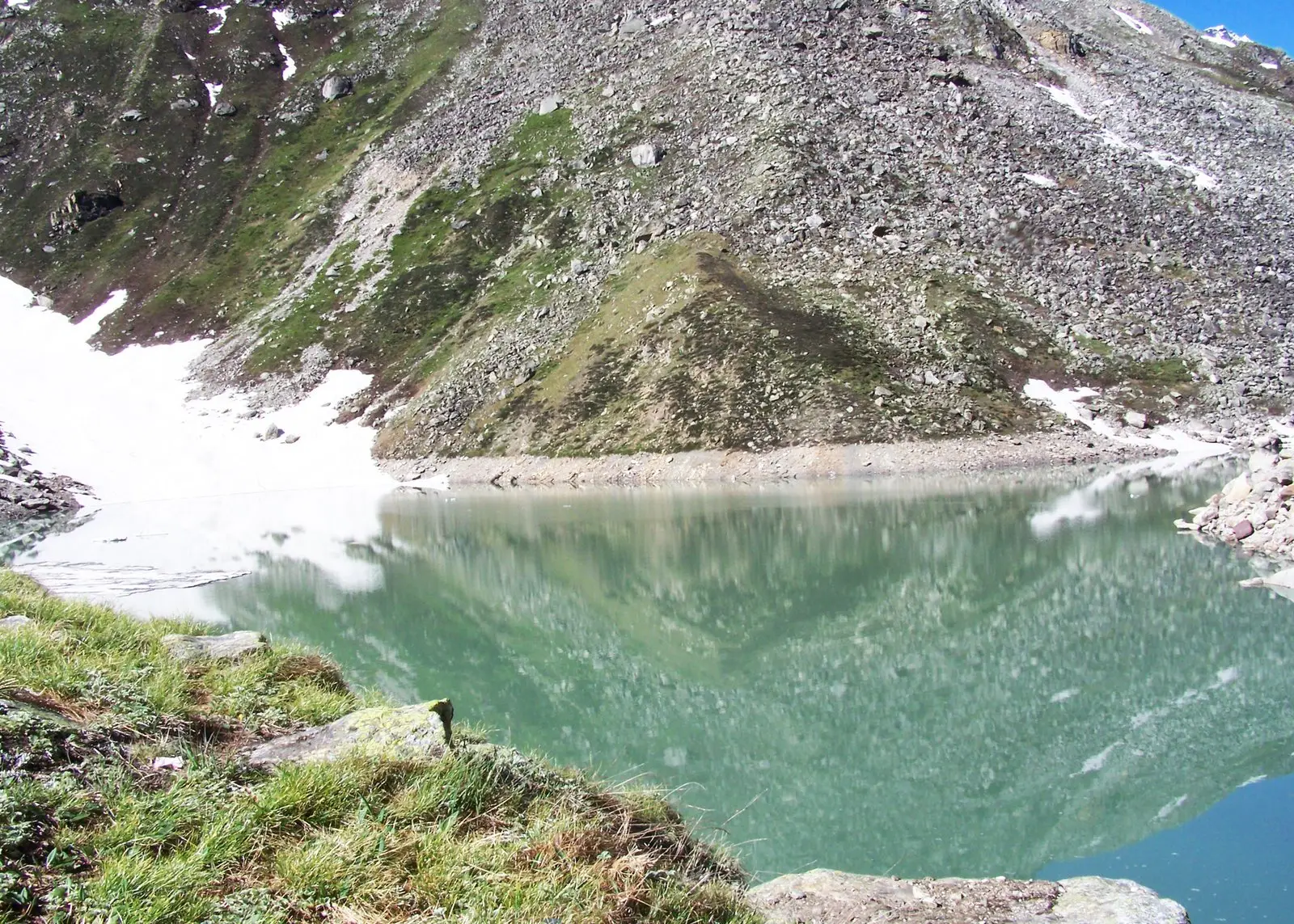 Green water of Satopanth Tal reflecting a scree slope, with snow patches on the far shore and grass at the near edge