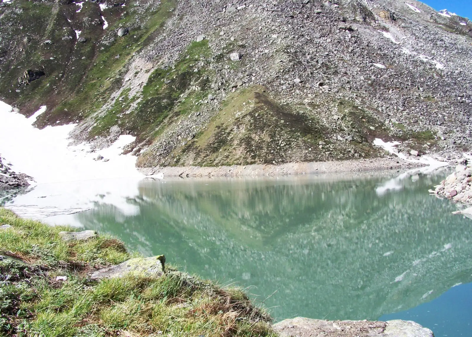 Green water of Satopanth Tal reflecting a scree slope, with snow patches on the far shore and grass at the near edge