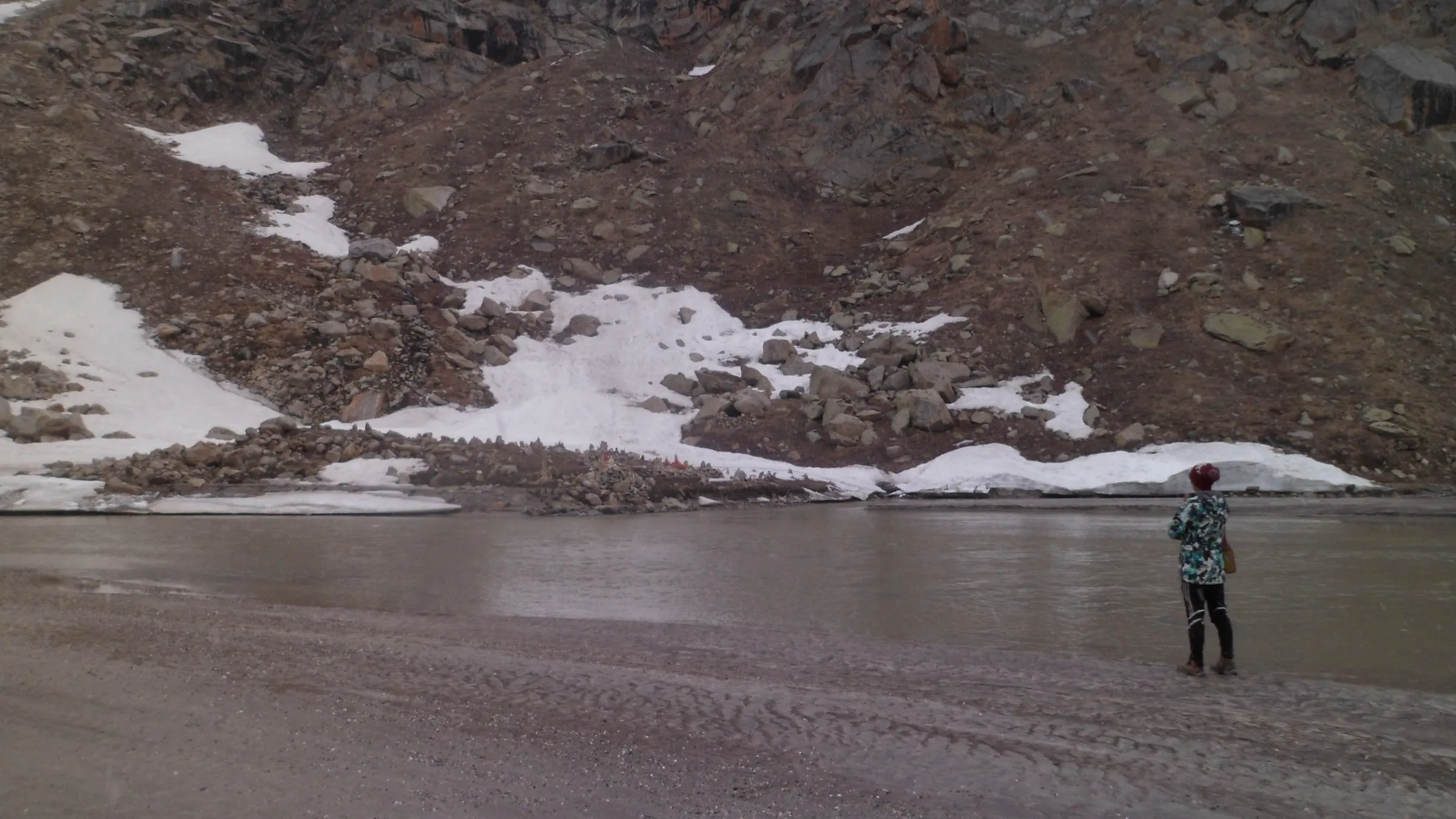 A trekker standing on the silty shore of Mantalai Lake below a rocky slope streaked with snow