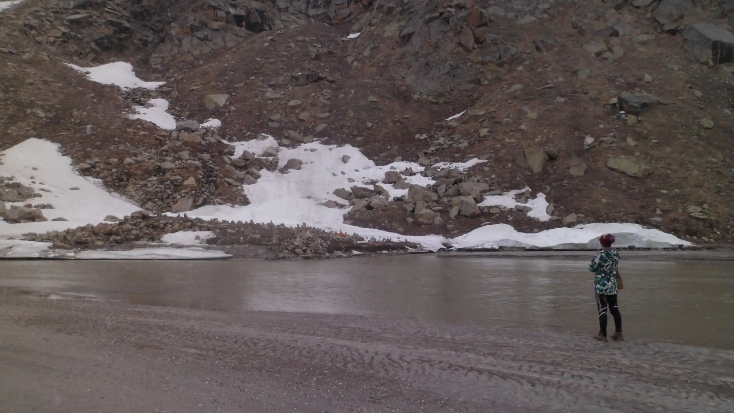 A trekker standing on the silty shore of Mantalai Lake below a rocky slope with patches of snow