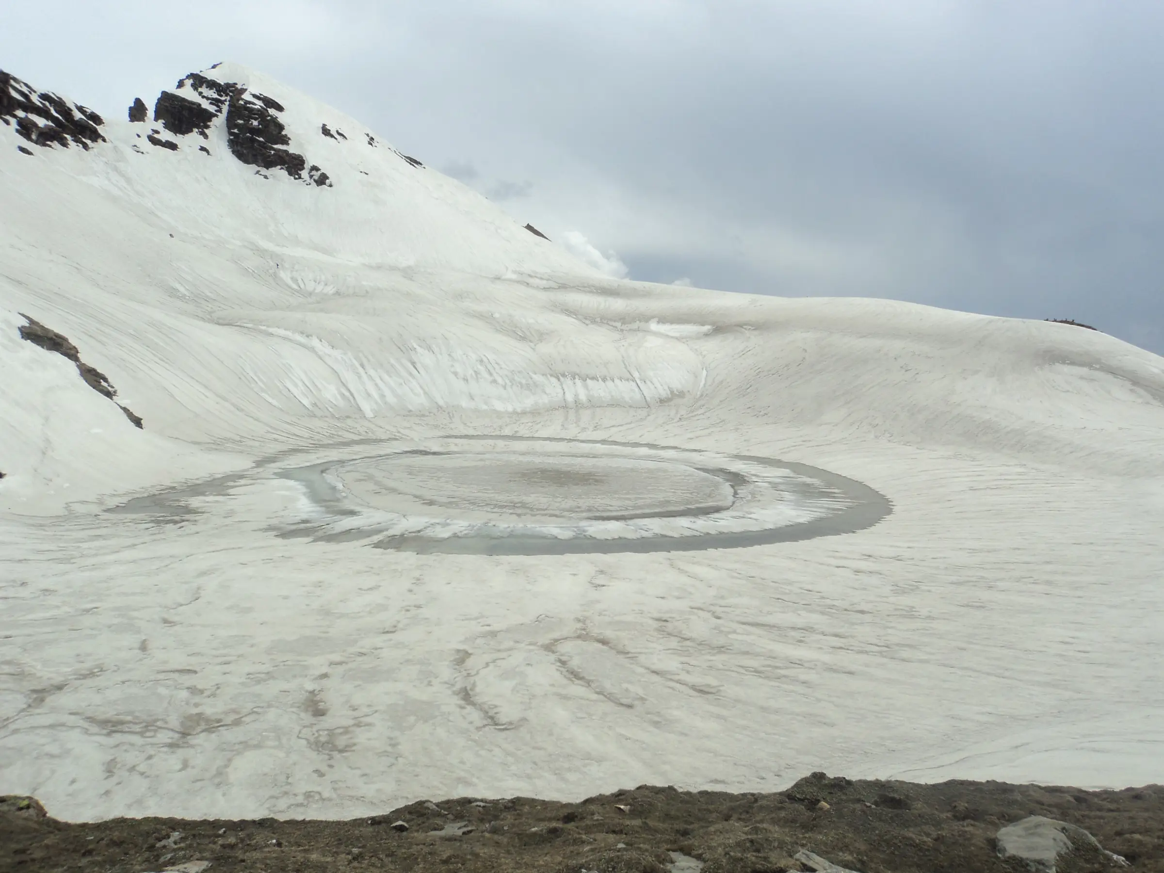 Partly frozen Bhrigu Lake ringed by thawing ice in a bowl of snow below a rocky snow-covered peak