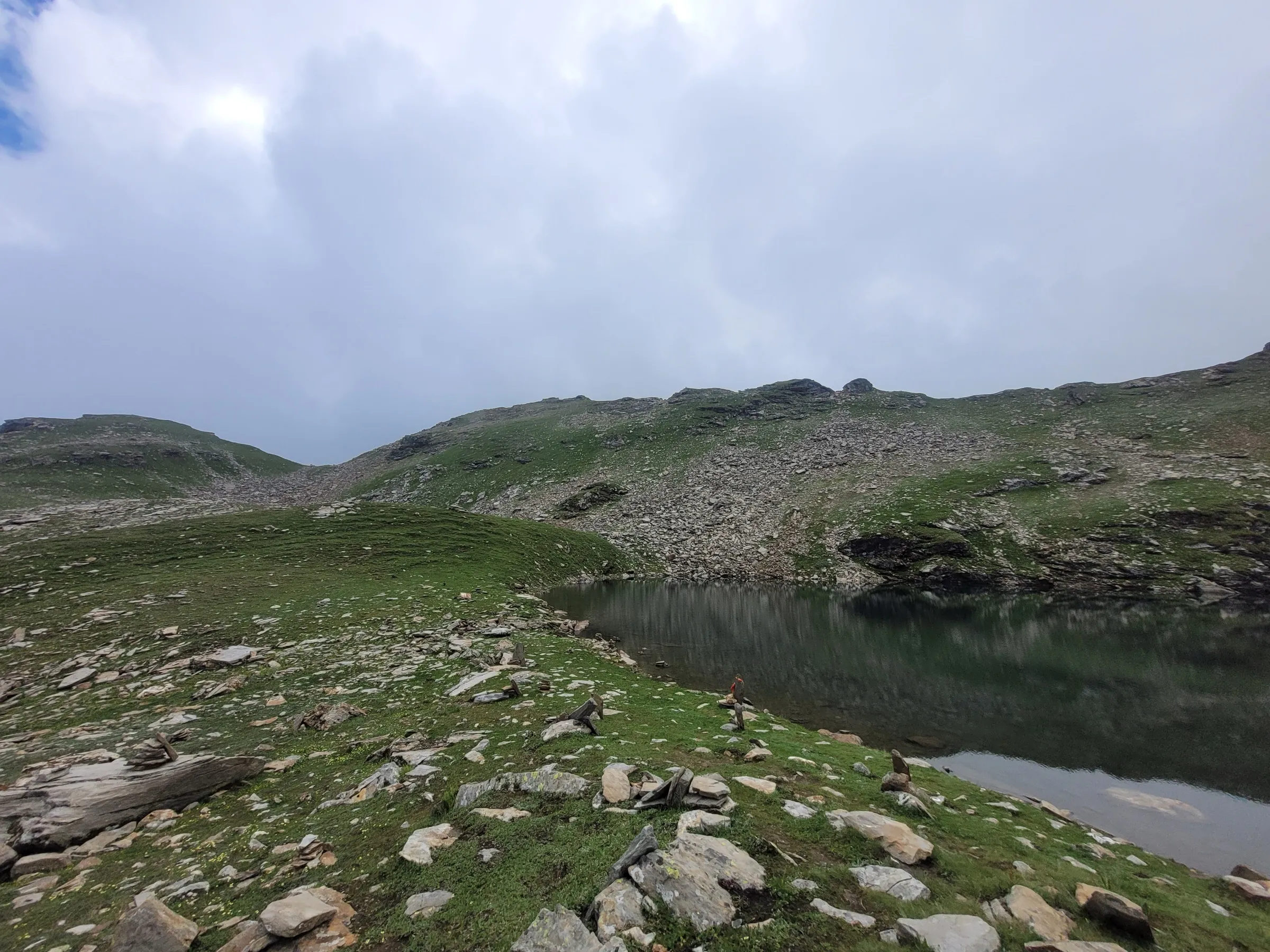 Bhrigu Lake in summer, a dark still lake beside green grassy slopes scattered with stones and small cairns under heavy cloud