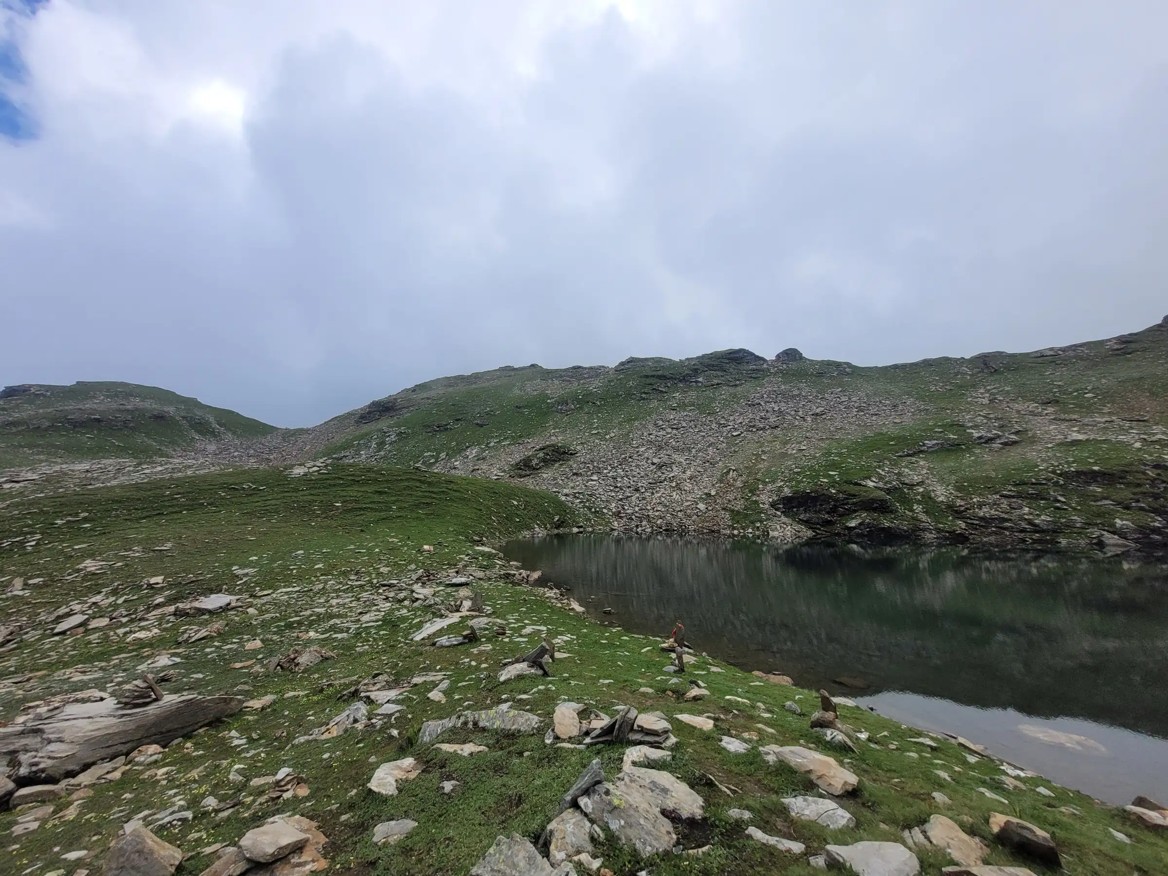 Bhrigu Lake in summer, a dark still lake beside green grassy slopes scattered with stones and small cairns under heavy cloud