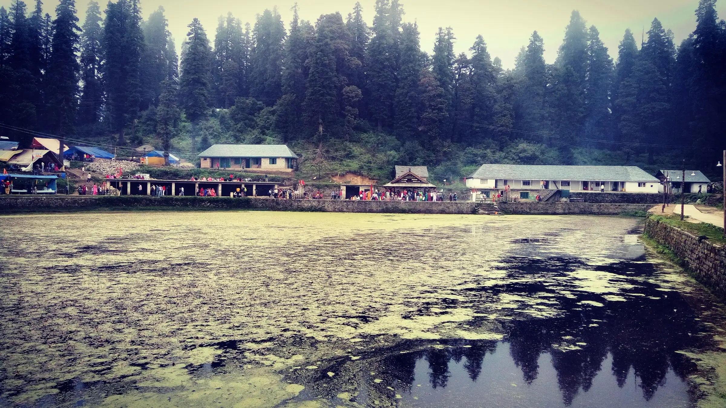 Kamrunag lake with the temple of Dev Kamrunag and pilgrim shelters on its far bank, under tall deodar forest