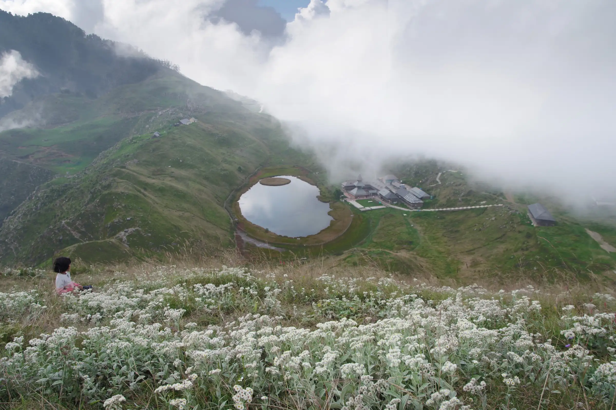 Prashar Lake with its round floating island and the temple buildings beside it, seen from a slope of white wildflowers under drifting cloud