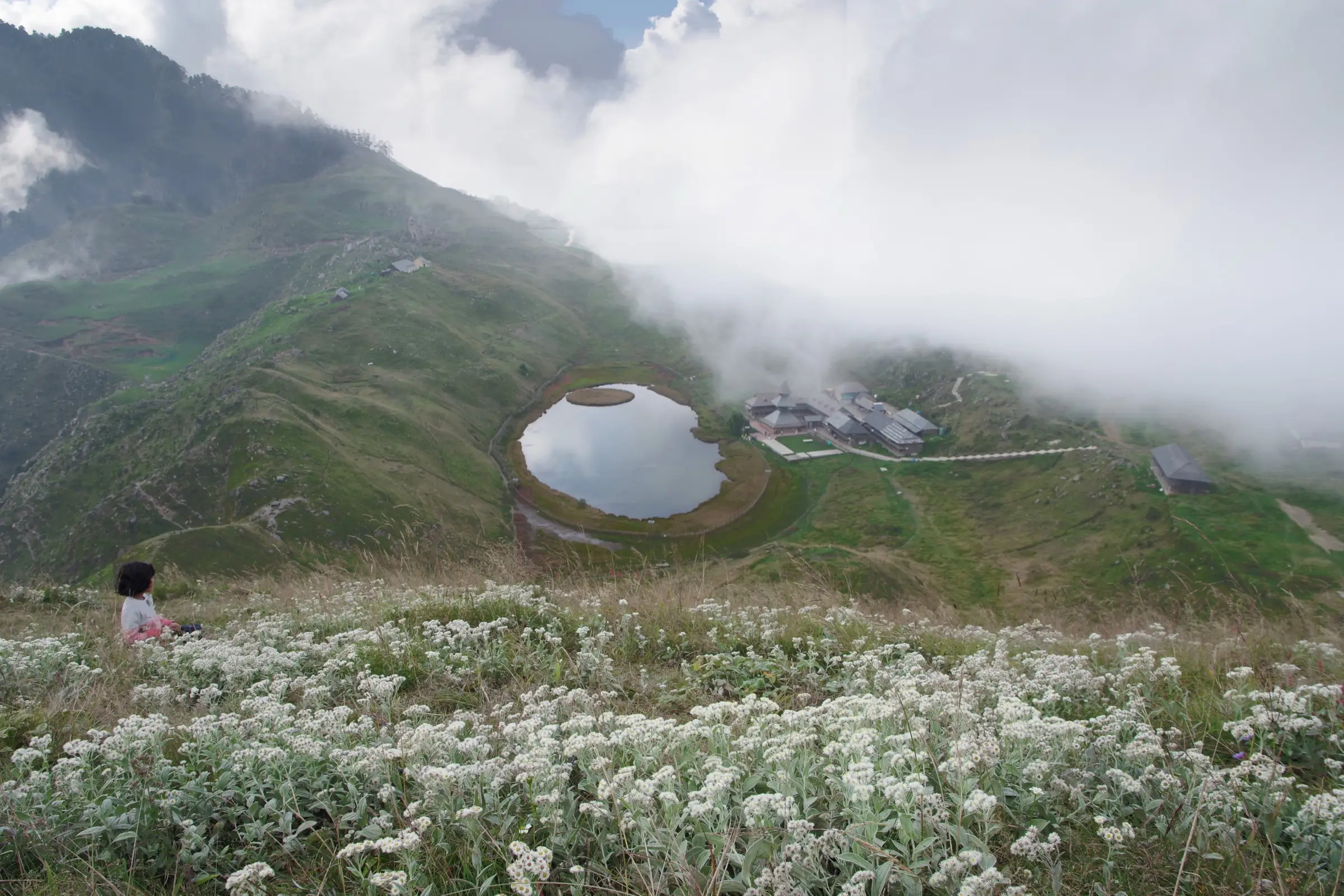 Prashar Lake with its round floating island and the temple buildings beside it, seen from a slope of white wildflowers under drifting cloud
