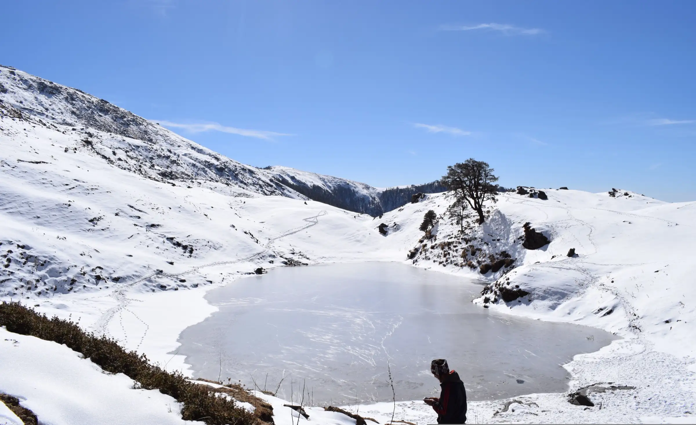 Frozen Brahmatal lake in a snowy bowl below a ridge, with a lone tree and a trekker in the foreground under a blue sky