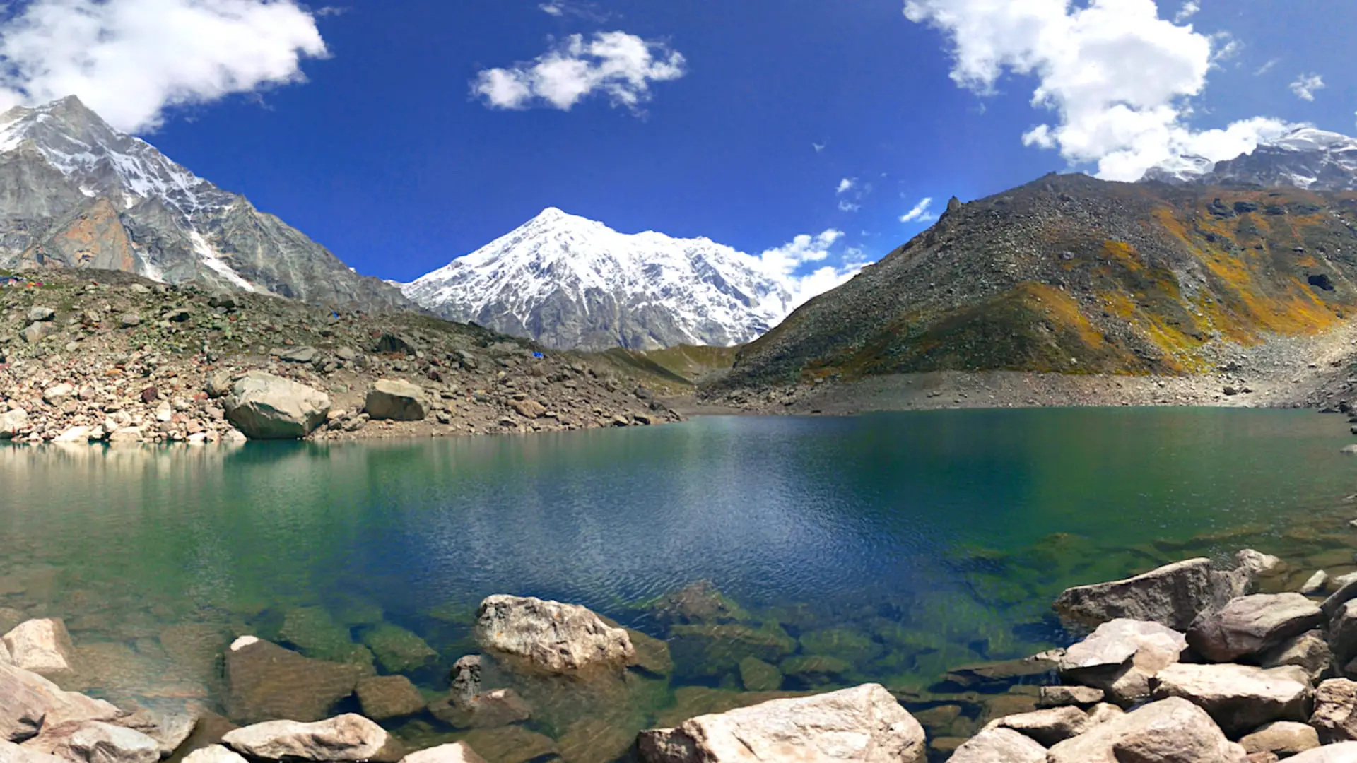 Emerald-green Satopanth Tal with boulders in the foreground, a snow-covered peak rising behind and moraine slopes on either side under a blue sky