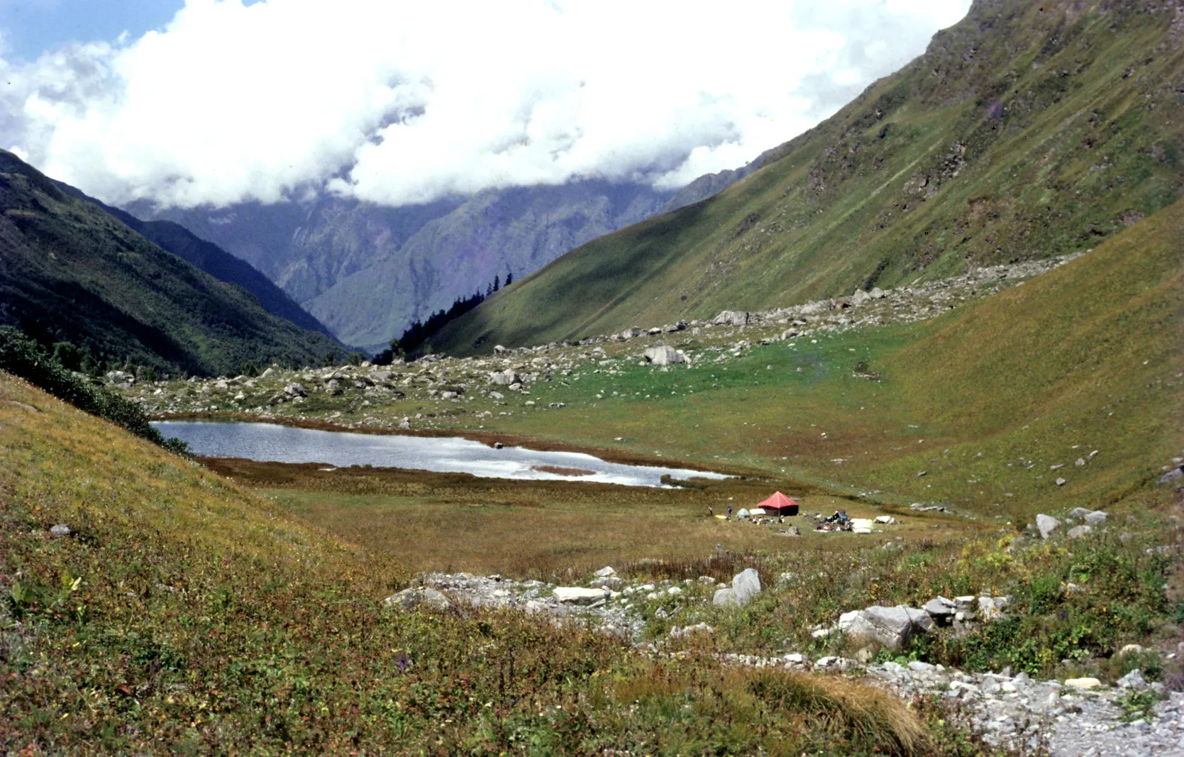 Ruinsara Tal, a small lake in a green meadow with a red tent beside it, under clouded mountains
