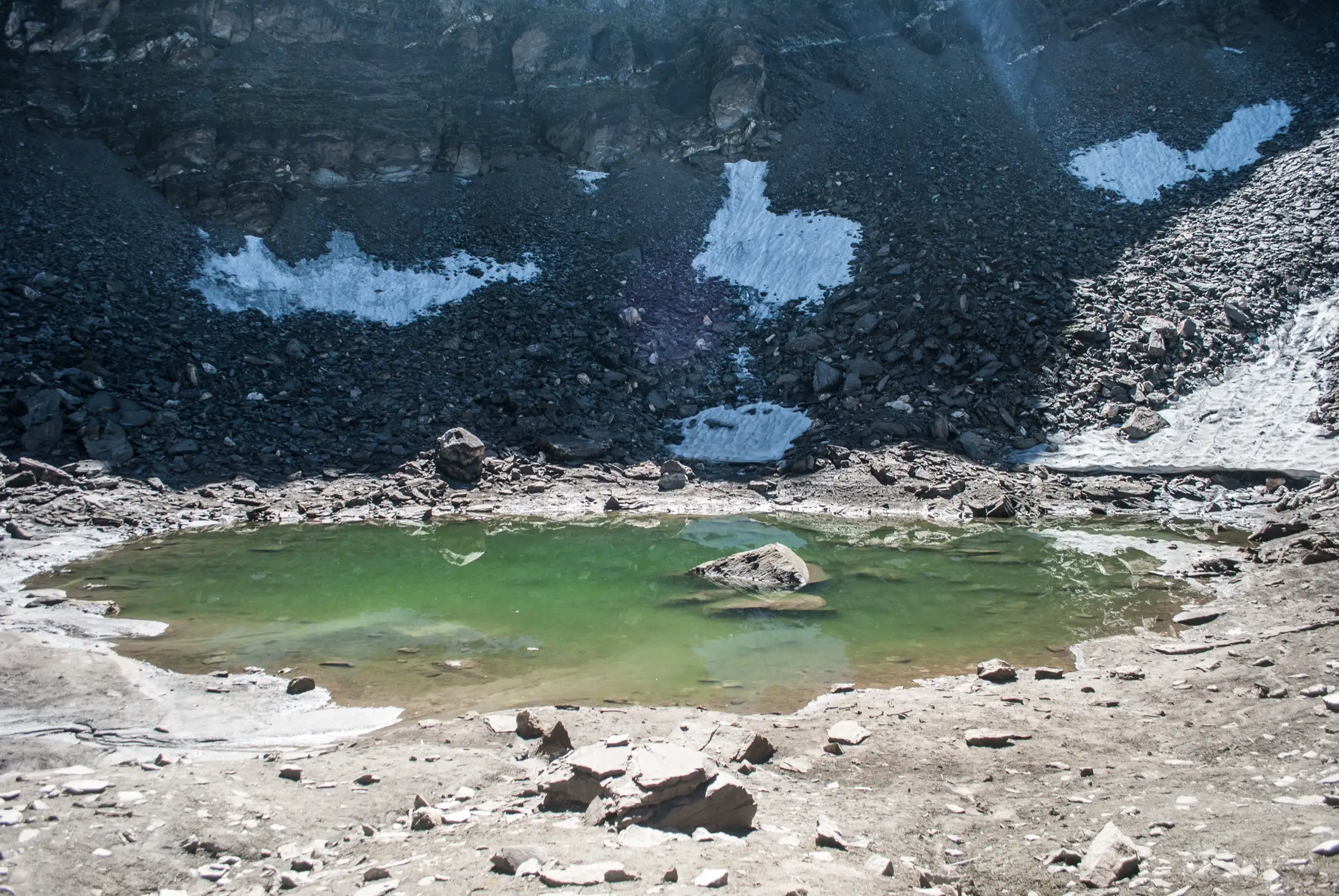 Small green glacial lake of Roopkund in a stony basin with patches of snow on the dark scree slopes above