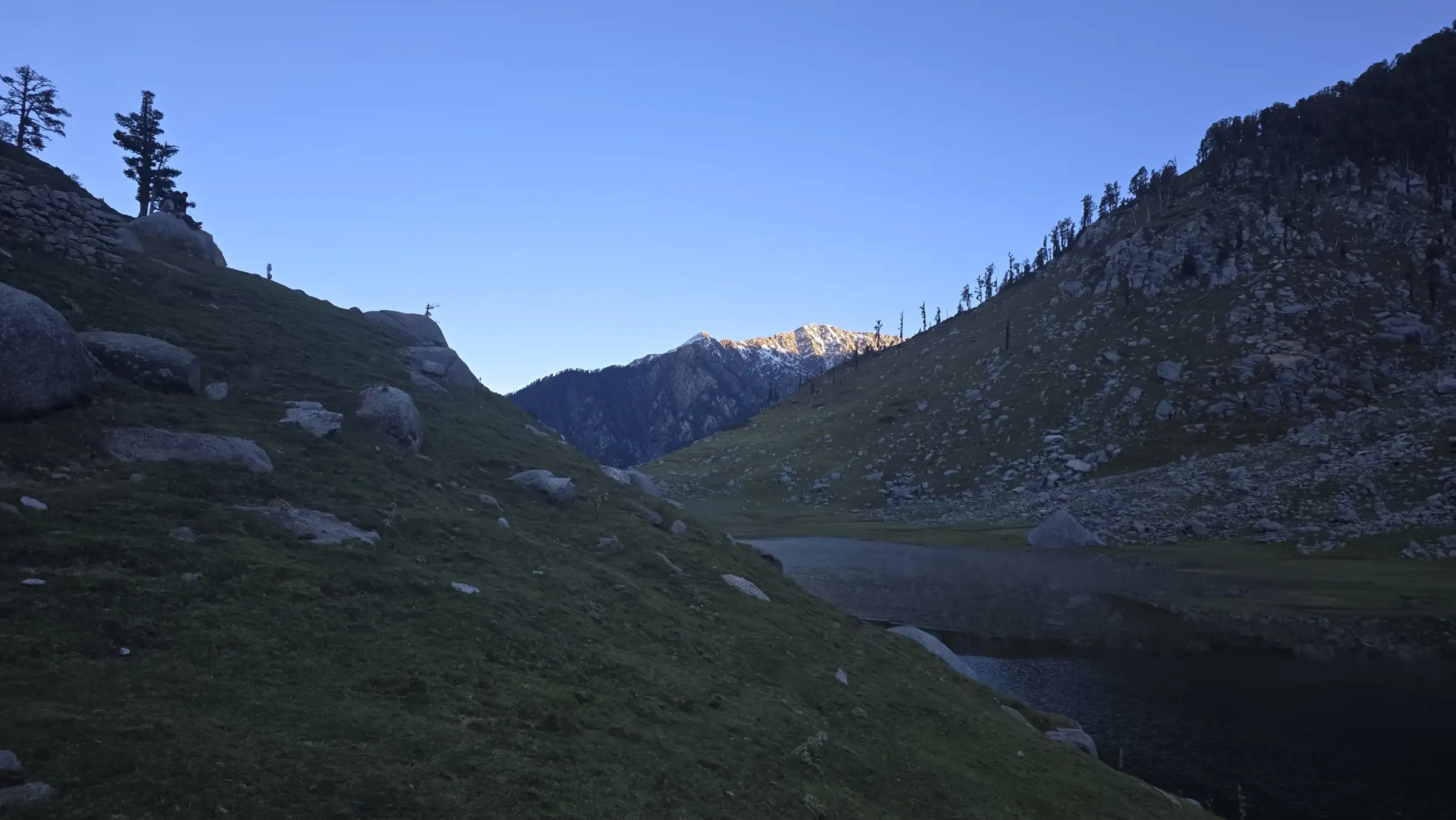 Kareri Lake at dusk in a grassy valley with a sunlit snow peak at the far end