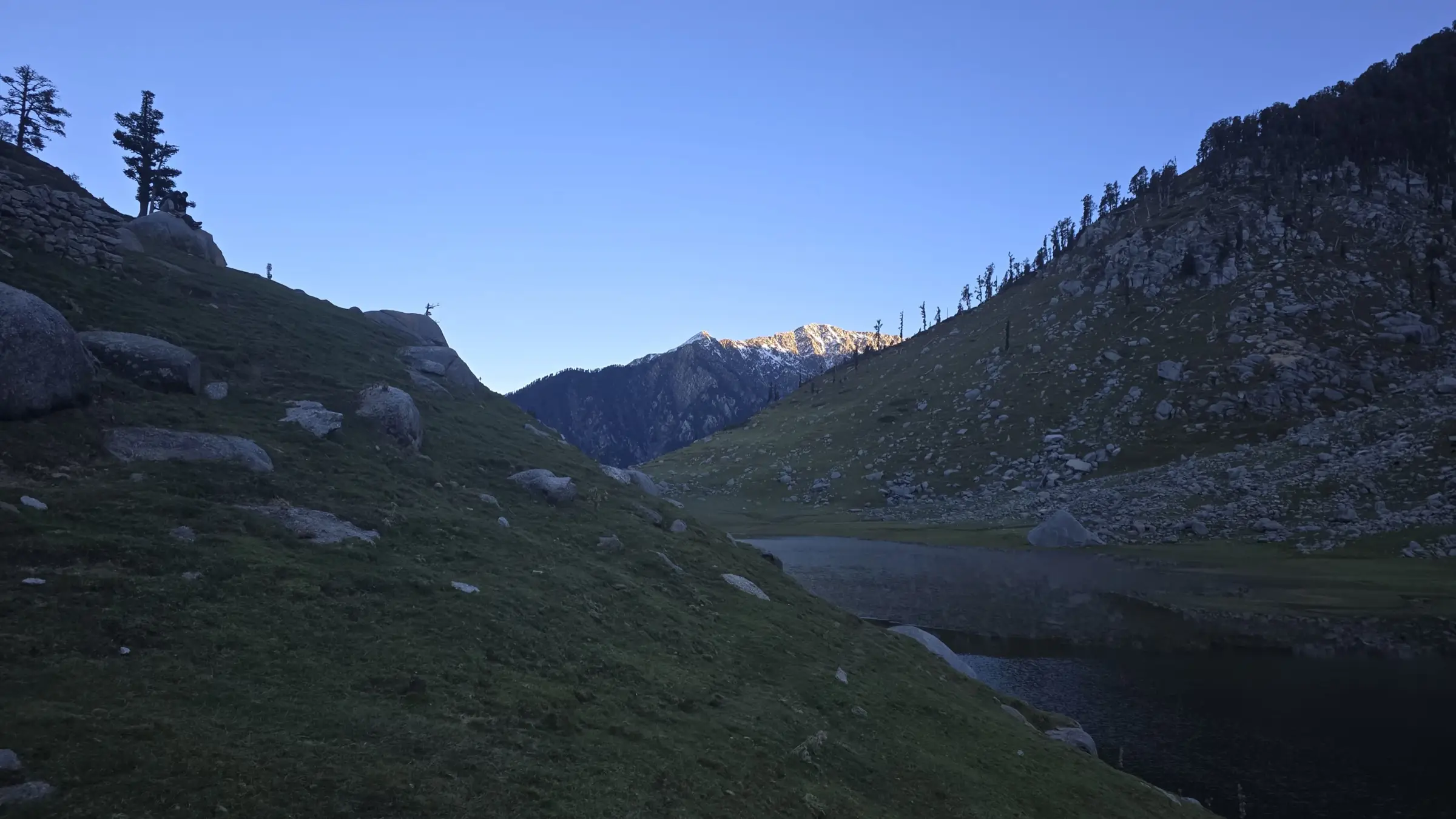 Kareri Lake at dusk in a grassy valley with a sunlit snow peak at the far end