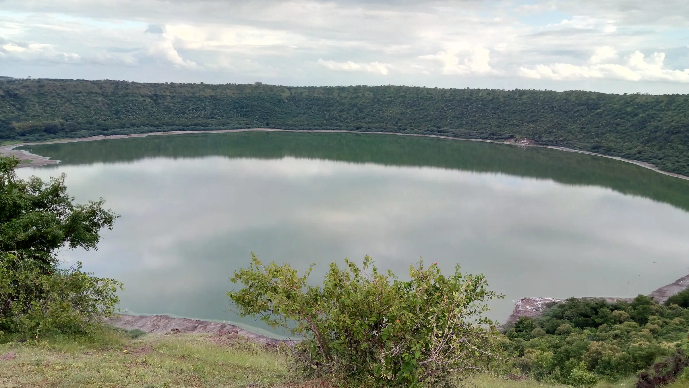 Lonar crater lake seen from the rim, its green water ringed by a forested crater wall under a cloudy sky