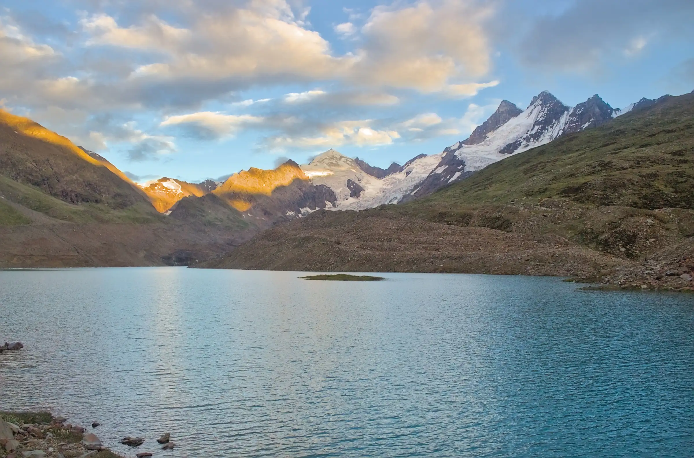 Blue Ghepan Lake with a small grassy island, below glaciated peaks lit by the evening sun
