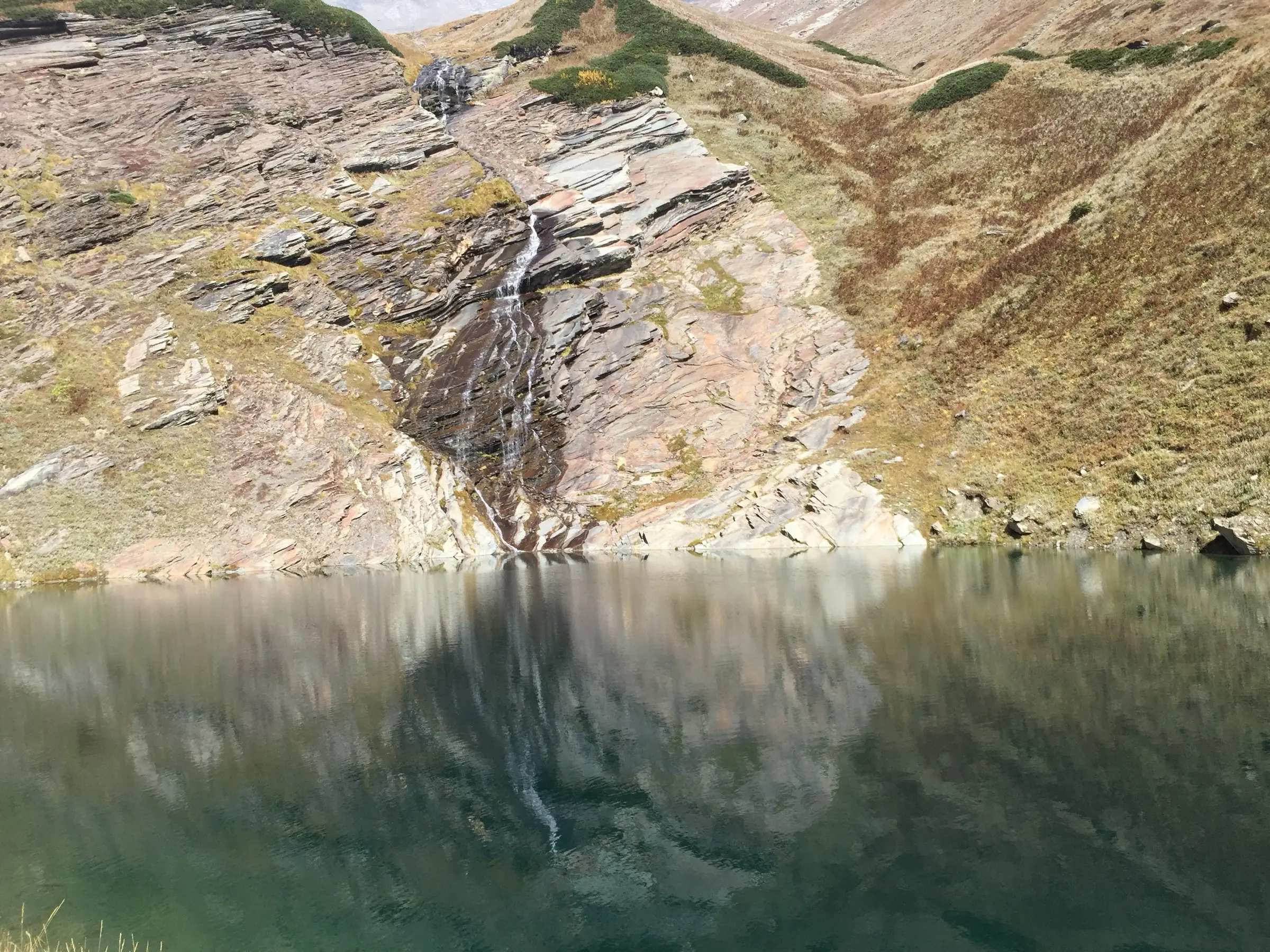 A thin waterfall running down striped rock into the still water of Beas Kund in August