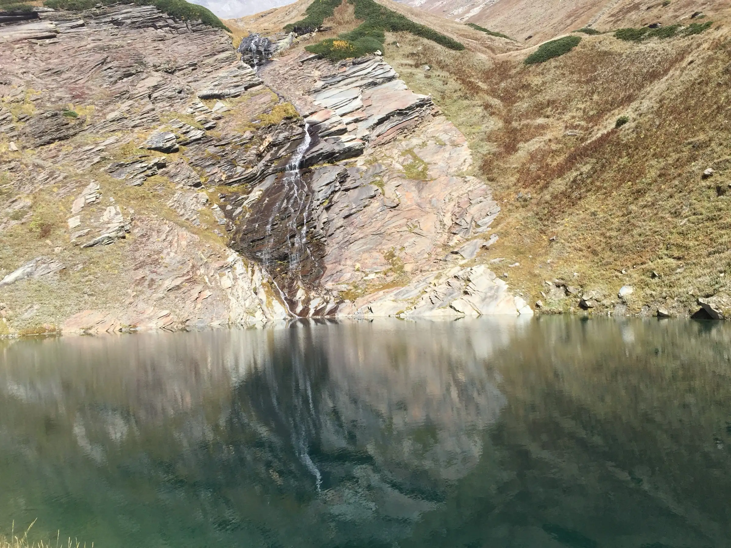A thin waterfall running down striped rock into the still water of Beas Kund in August