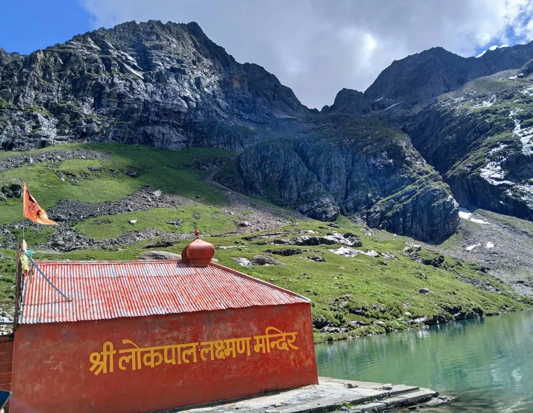 Red-roofed Shri Lokpal Lakshman Mandir with its saffron flag beside the green Hemkund lake and rocky mountains