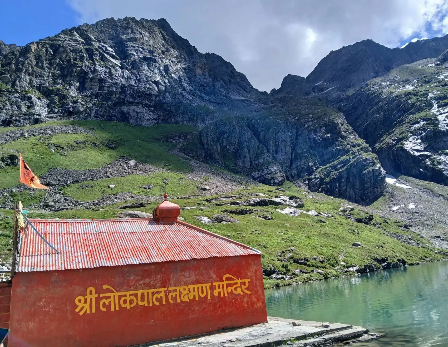 Red-roofed Shri Lokpal Lakshman Mandir with its saffron flag beside the green Hemkund lake and rocky mountains