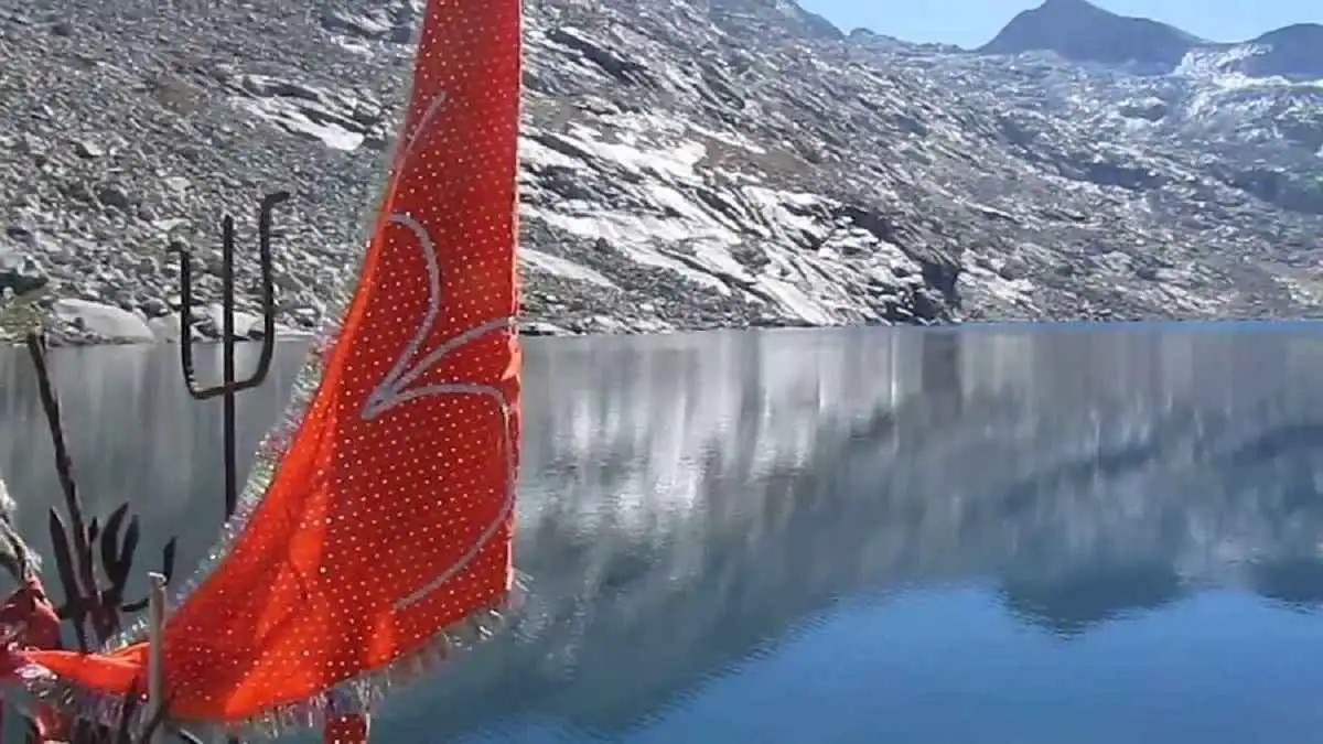 Still water of Lam Dal reflecting the rocky slopes, with a trident and a red flag offered on the shore