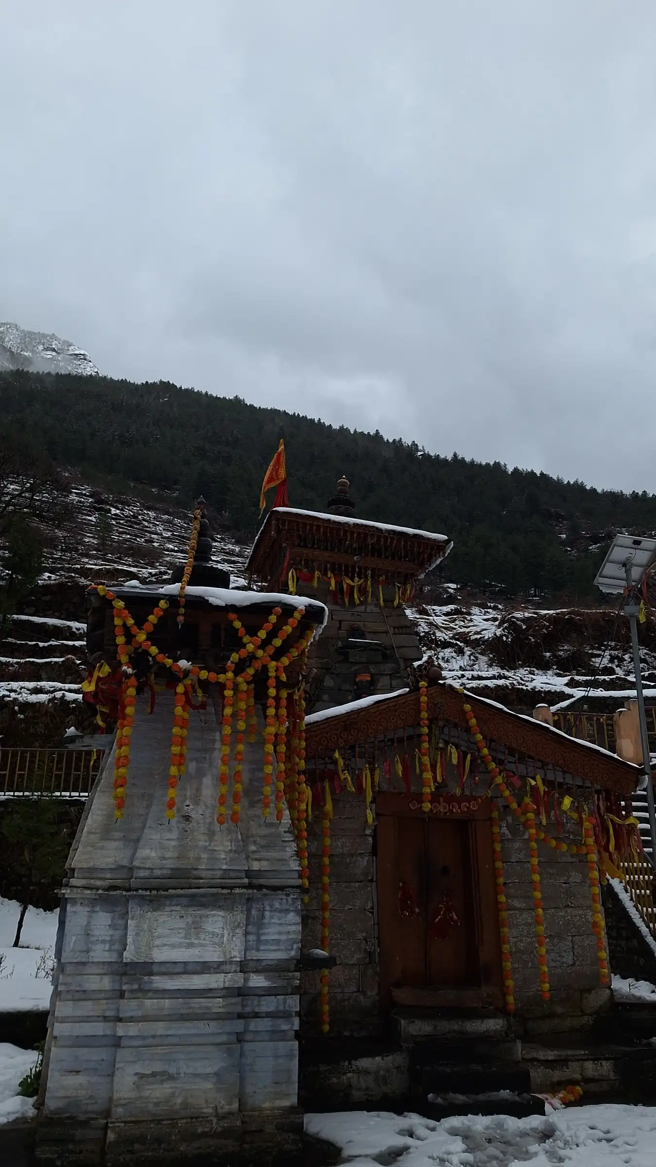 Nanda Devi temple in Lata village decorated with marigold garlands and a saffron flag, with fresh snow on the ground and forest behind