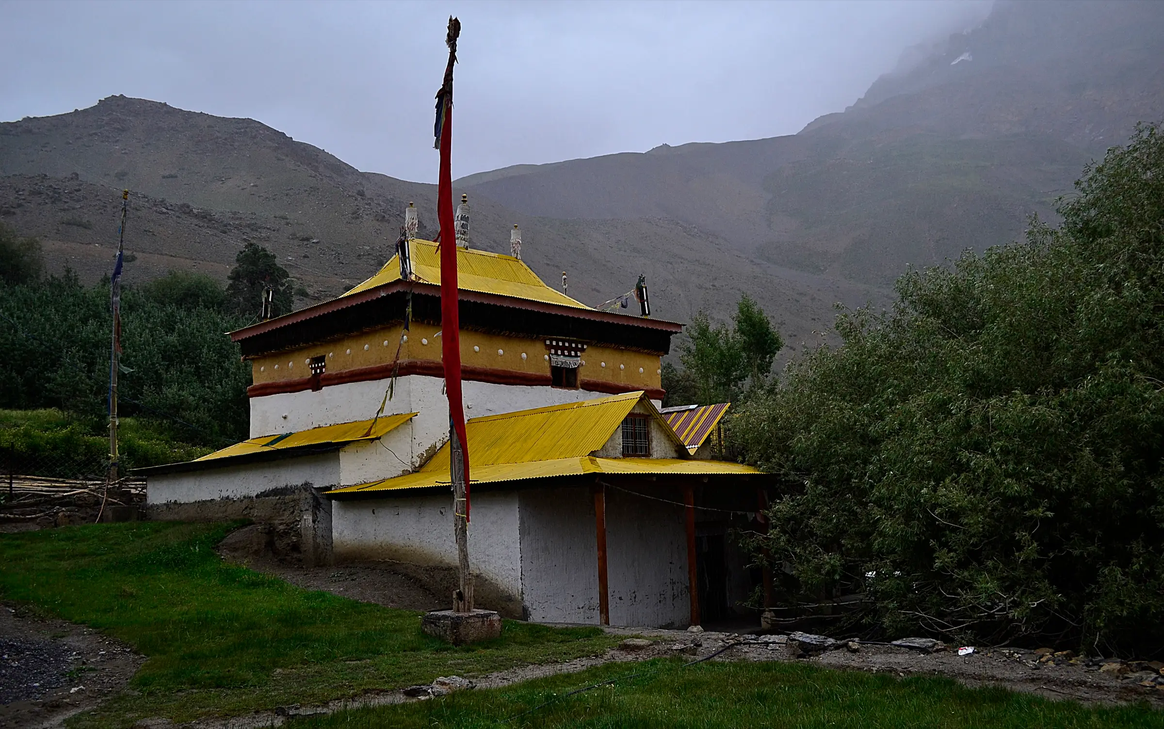 The yellow-roofed Lhalung (Serkhang) monastery among green trees and fields below bare grey mountains, on a cloudy day