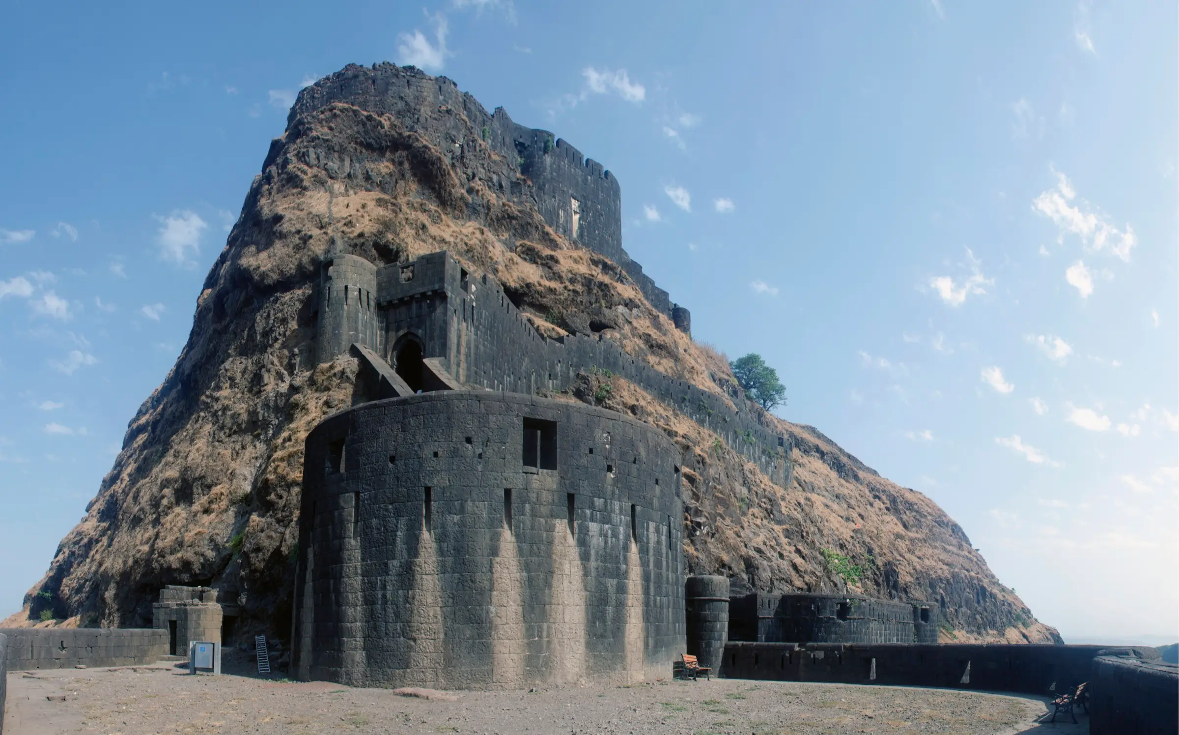 Massive round black-stone bastion and gateway walls of Lohagad Fort climbing up a dry, grassy rock face under a blue sky