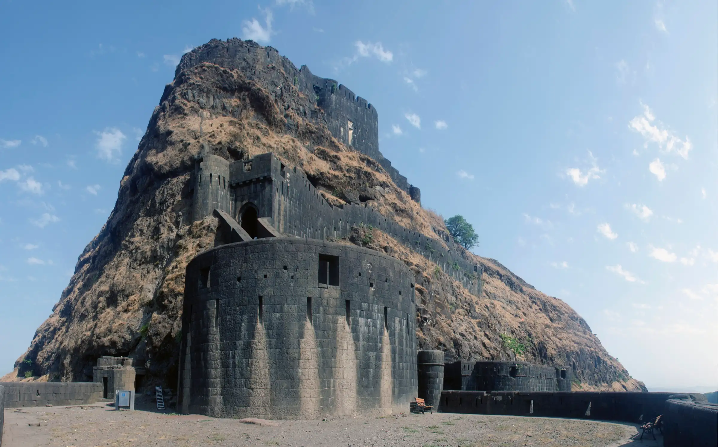 Massive round black-stone bastion and gateway walls of Lohagad Fort climbing up a dry, grassy rock face under a blue sky