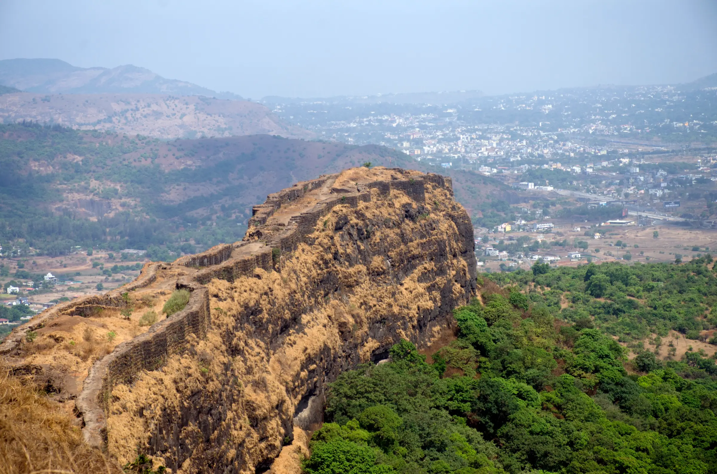 The walled Vinchu Kata spur of Lohagad stretching out above forested slopes, with Lonavala's buildings in the hazy valley behind