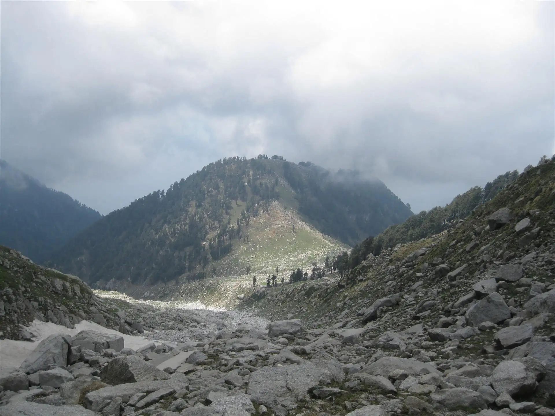Looking down the rocky trail at the green Laka Got meadow and forested ridge under monsoon clouds