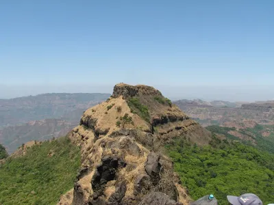 The rocky Madhu peak on a narrow forested ridge, seen from the top of Makrandgad fort under a hazy blue sky