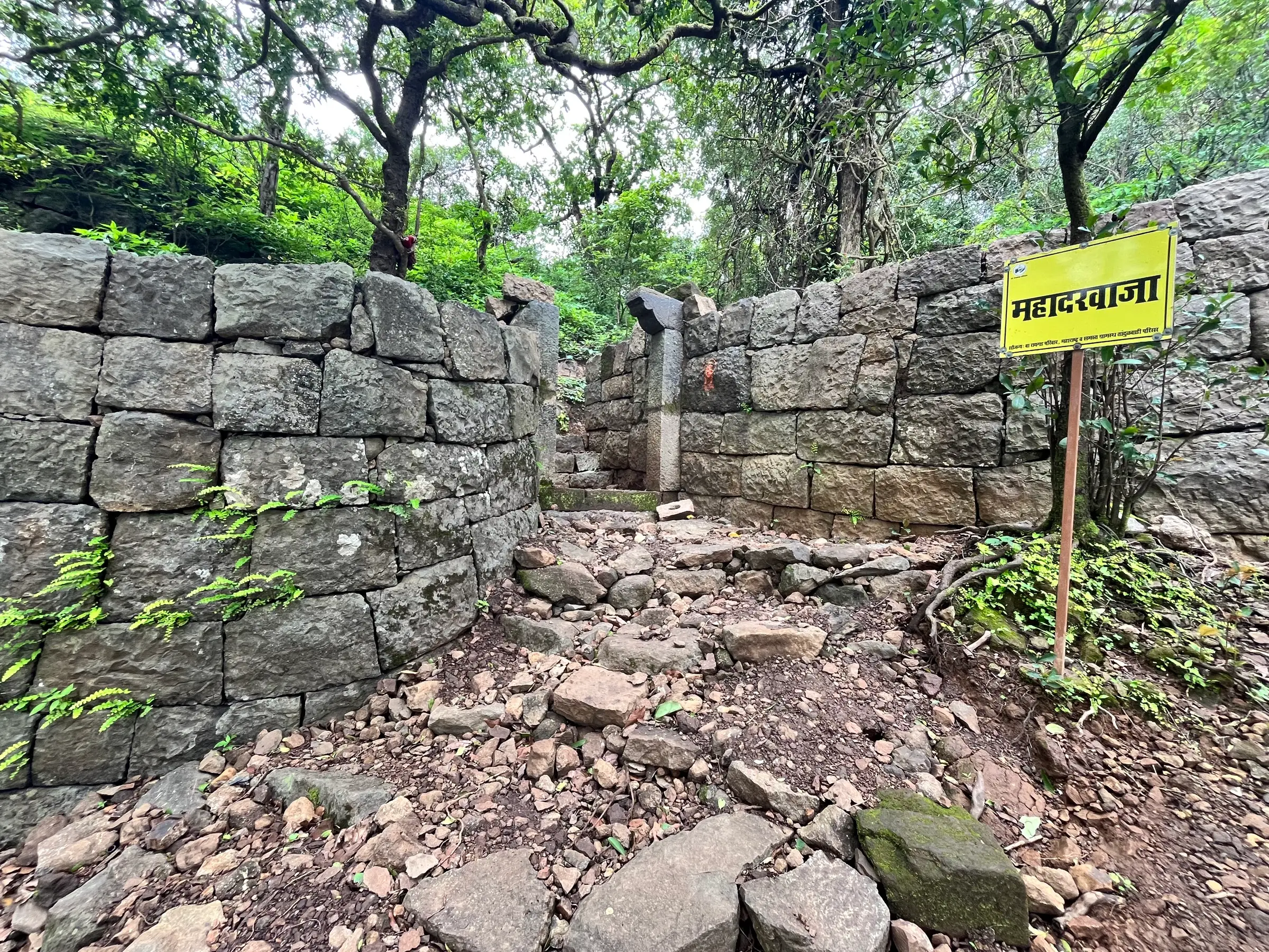 Ruined dry-stone walls and a rocky path at the Maha Darwaja of Tandulwadi Fort, with a yellow signboard