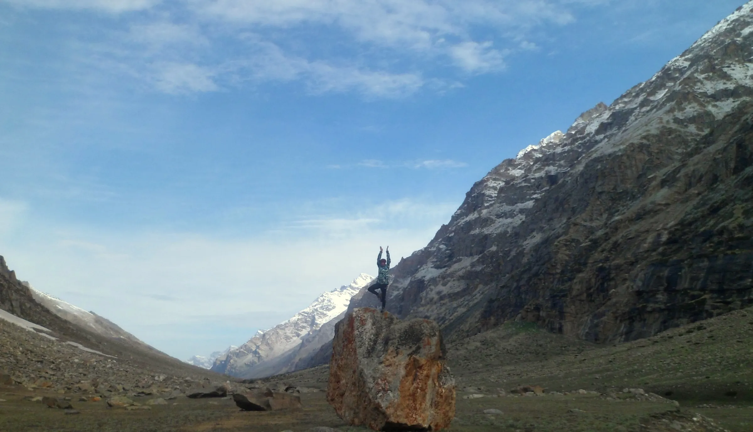 A trekker balancing on a boulder in the wide Parvati valley near Mantalai, with snow-streaked peaks under a blue sky