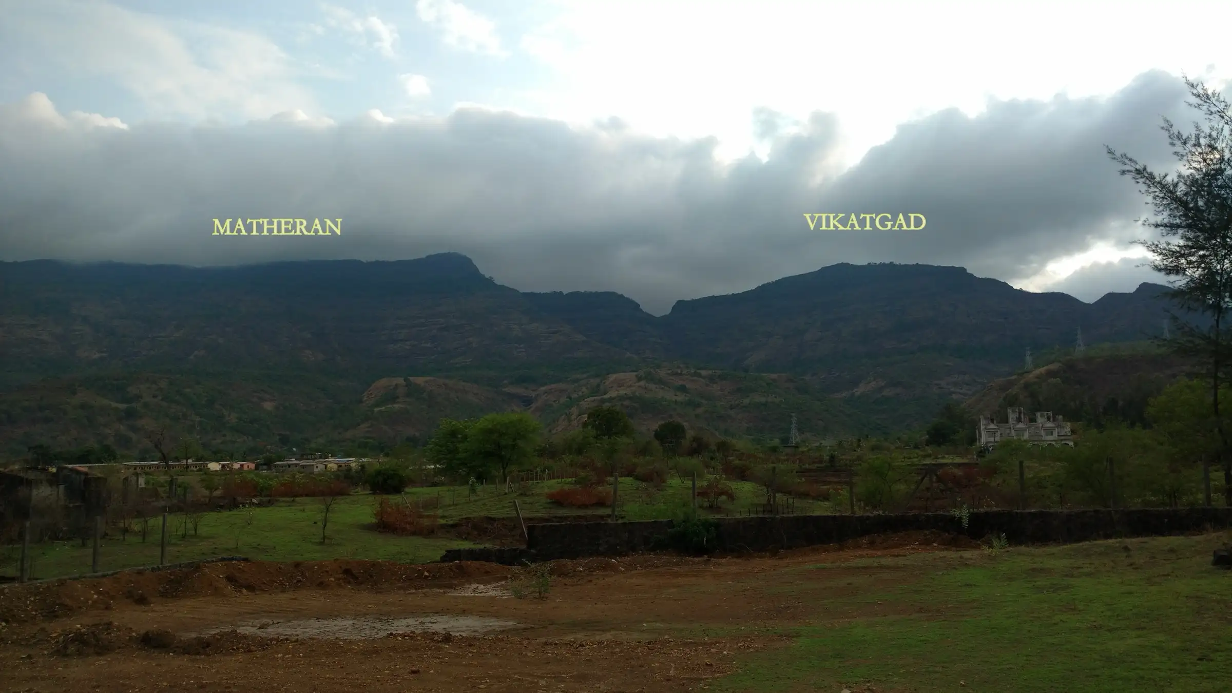 The Matheran plateau and the Vikatgad hill side by side under monsoon clouds, labelled, seen from the plains near Neral