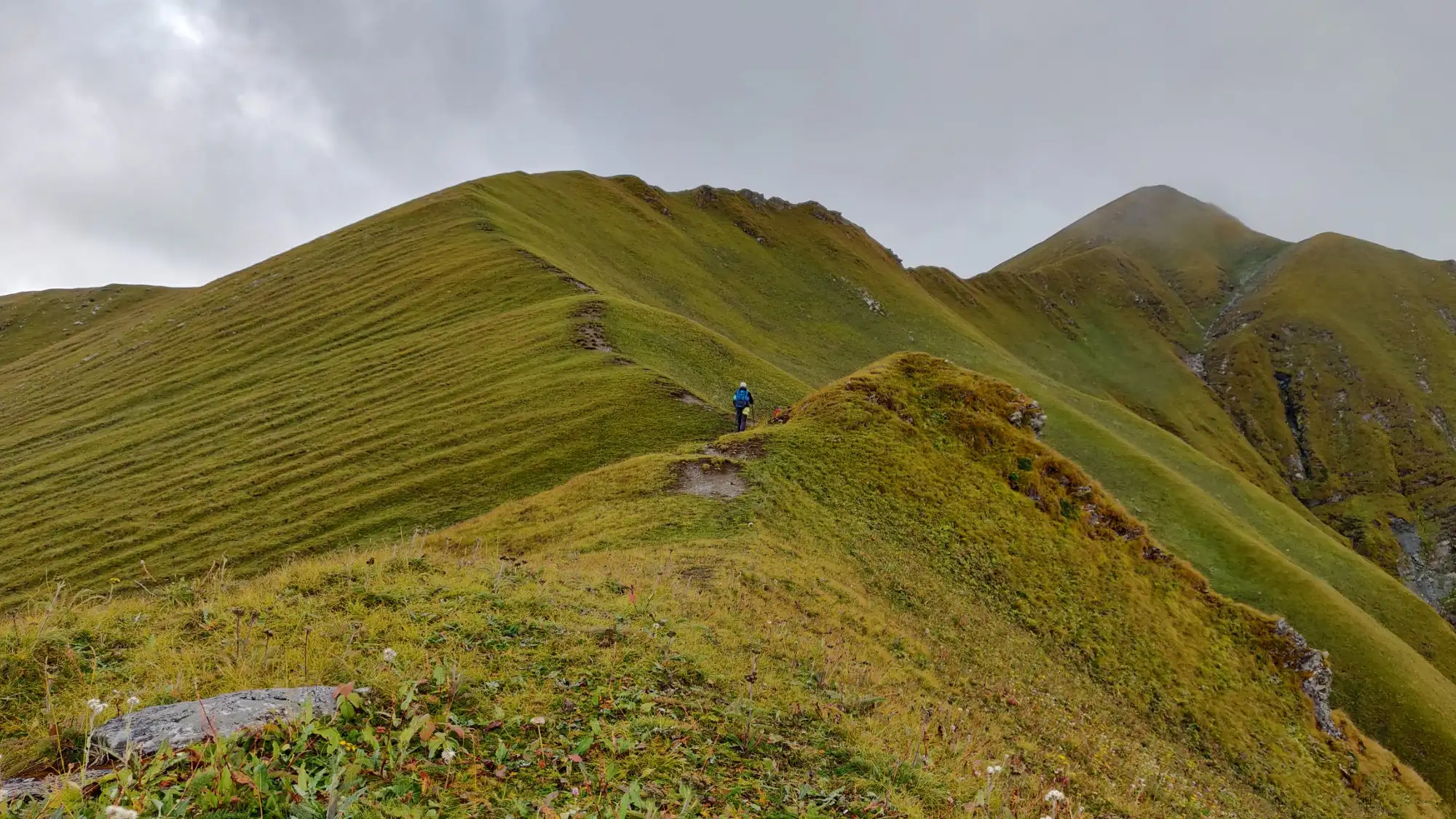 A trekker on a narrow grassy ridge in the Gidara Bugyal meadows under low grey cloud