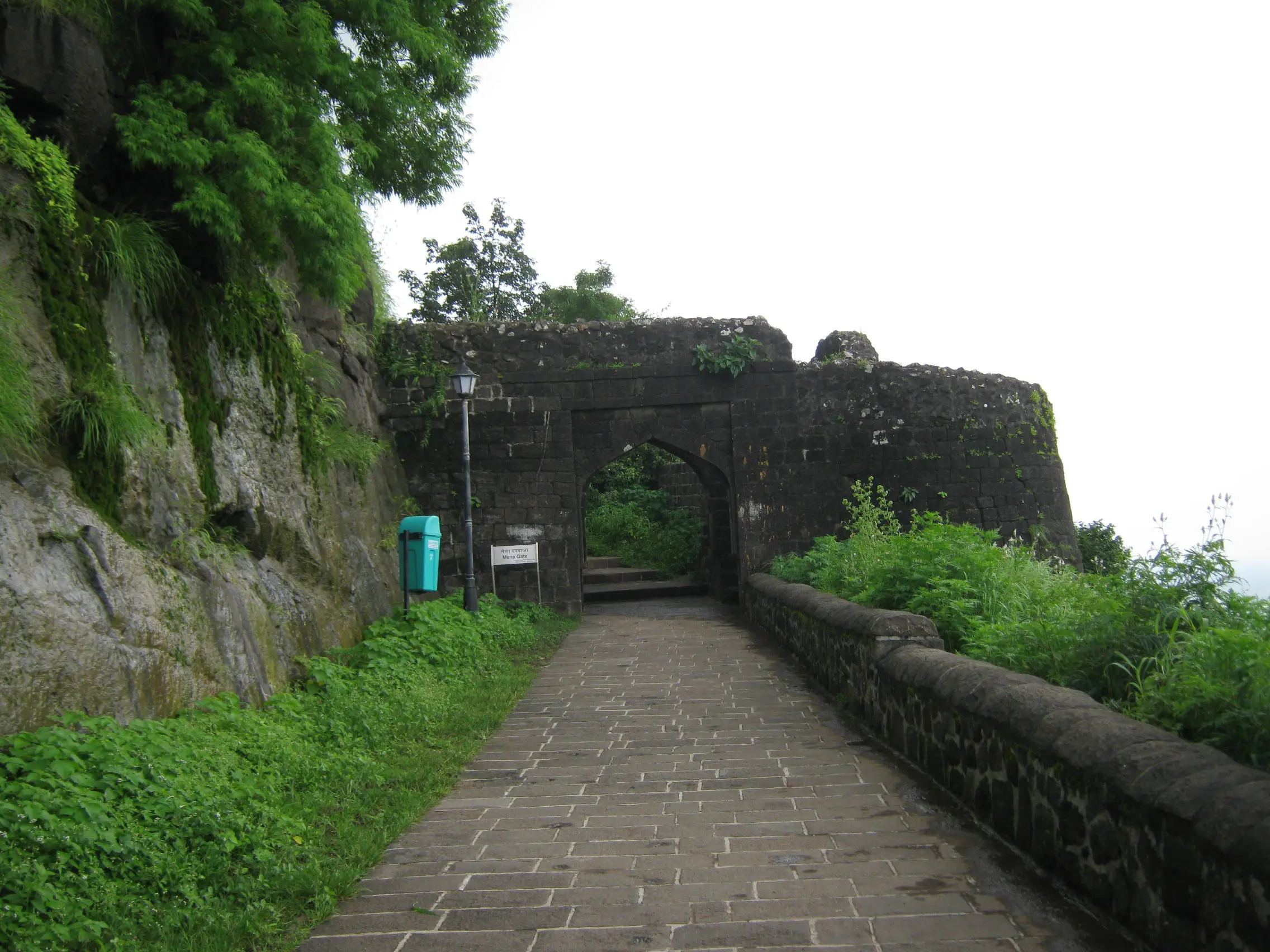Paved path between a rock face and a parapet wall leading to the Mena gate of Shivneri Fort in monsoon greenery