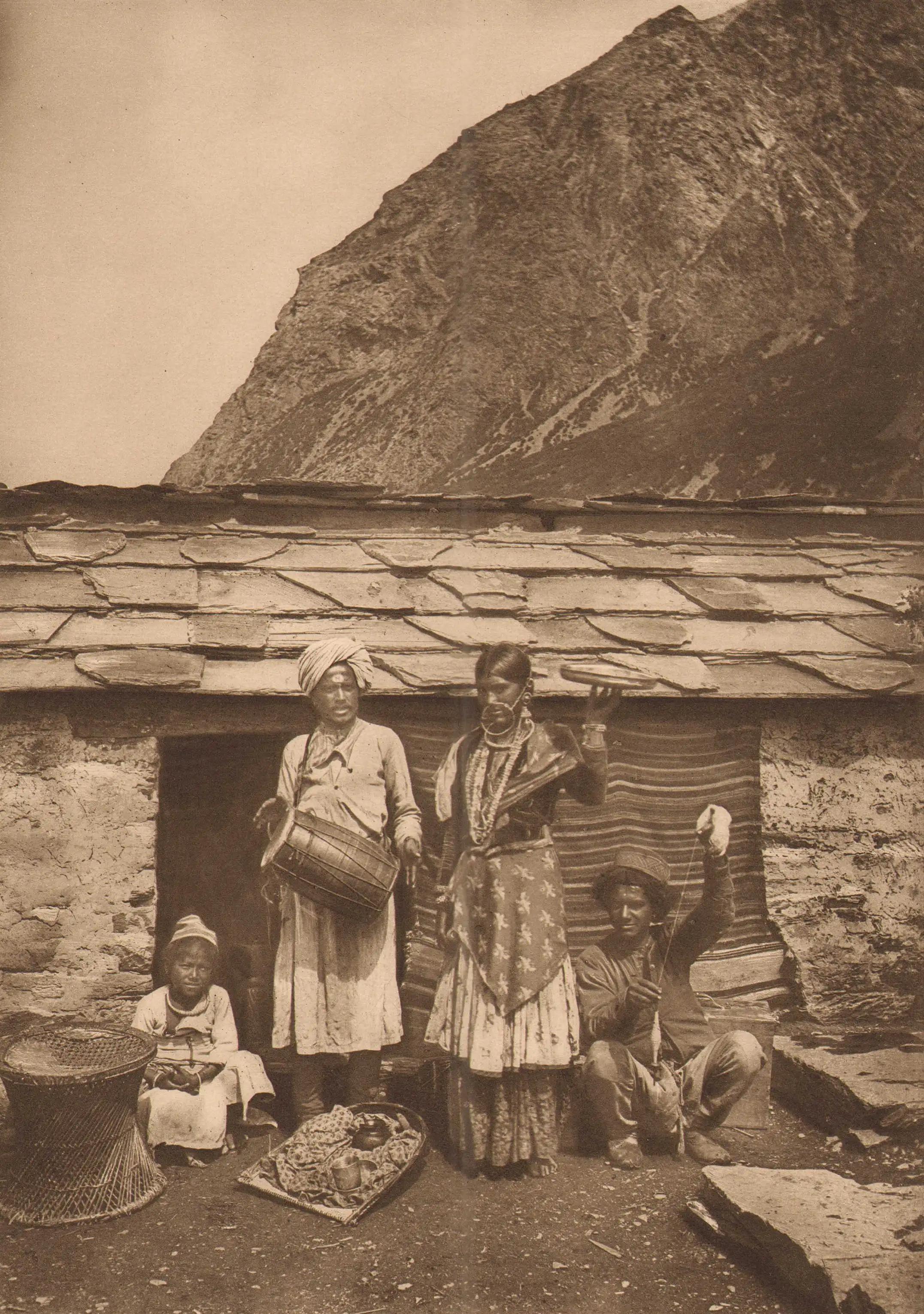 Old sepia photograph of a Johari family with a drum outside a slate-roofed stone house in Milam