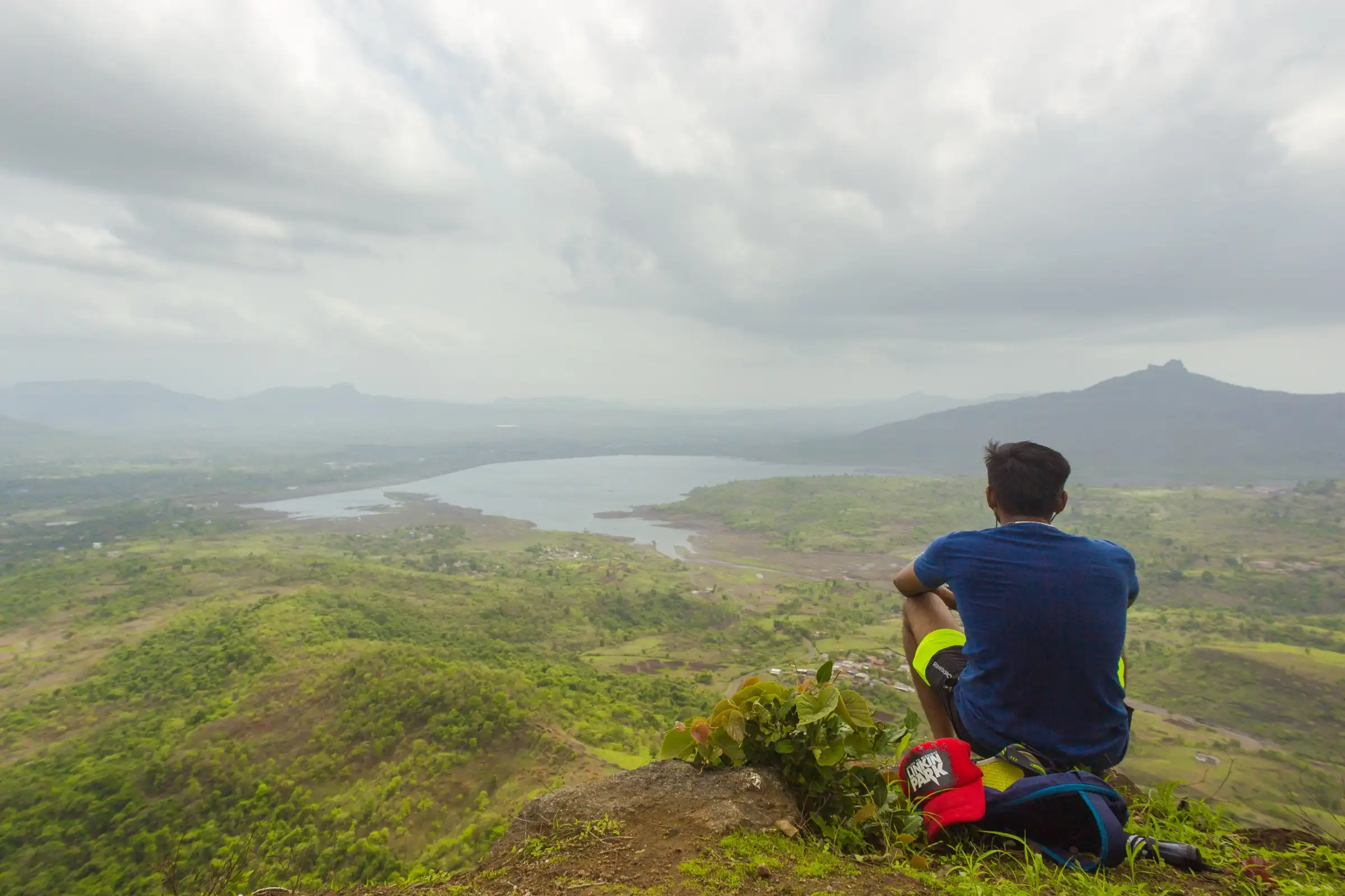 A trekker sitting on the edge of Sondai Fort looking over the Morbe Dam reservoir and green valley under monsoon clouds