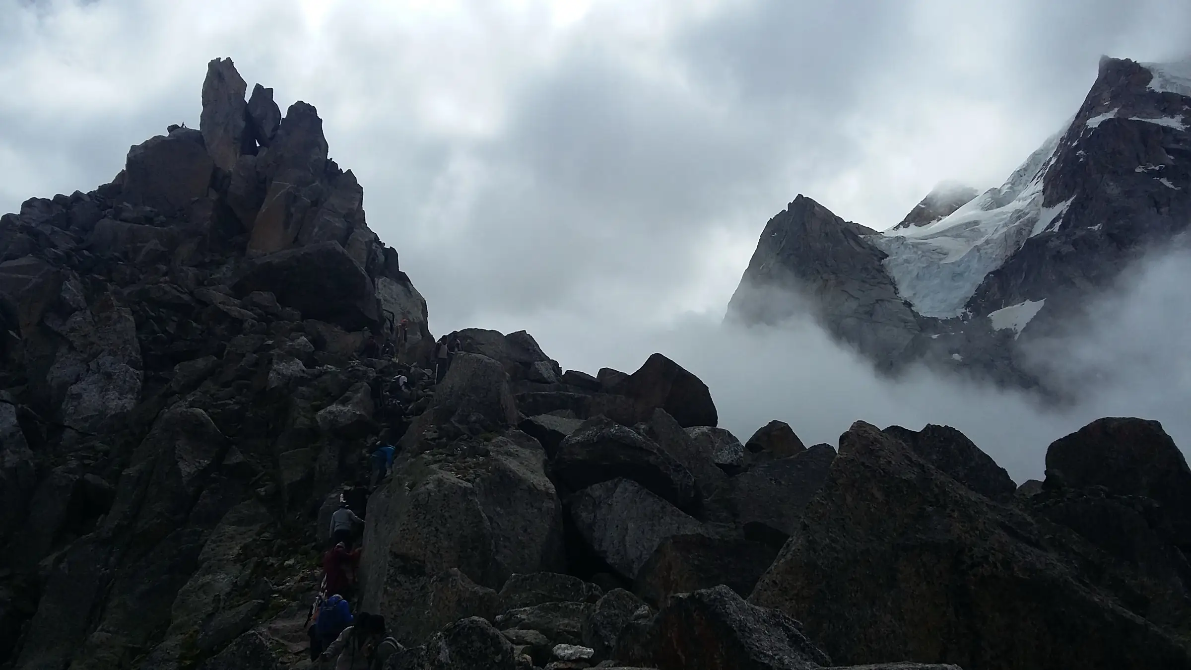 Pilgrims climbing between dark boulders in mist during the Kinner Kailash Yatra, with a glacier-covered peak to the right