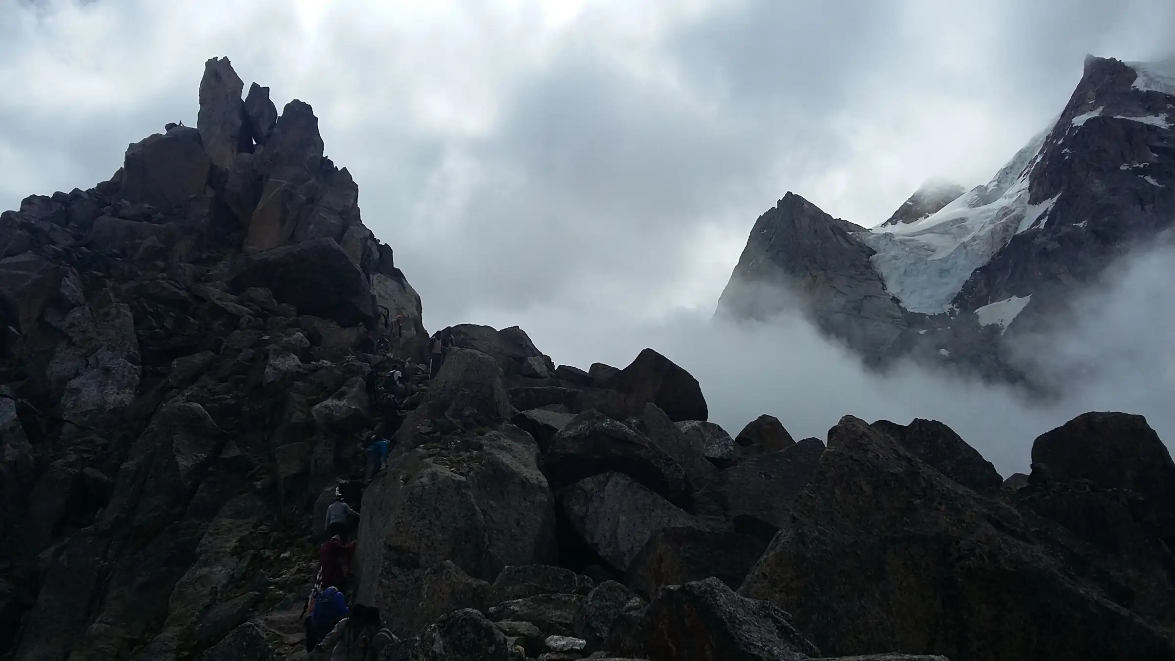 Pilgrims climbing between dark boulders in mist during the Kinner Kailash Yatra, with a glacier-covered peak to the right