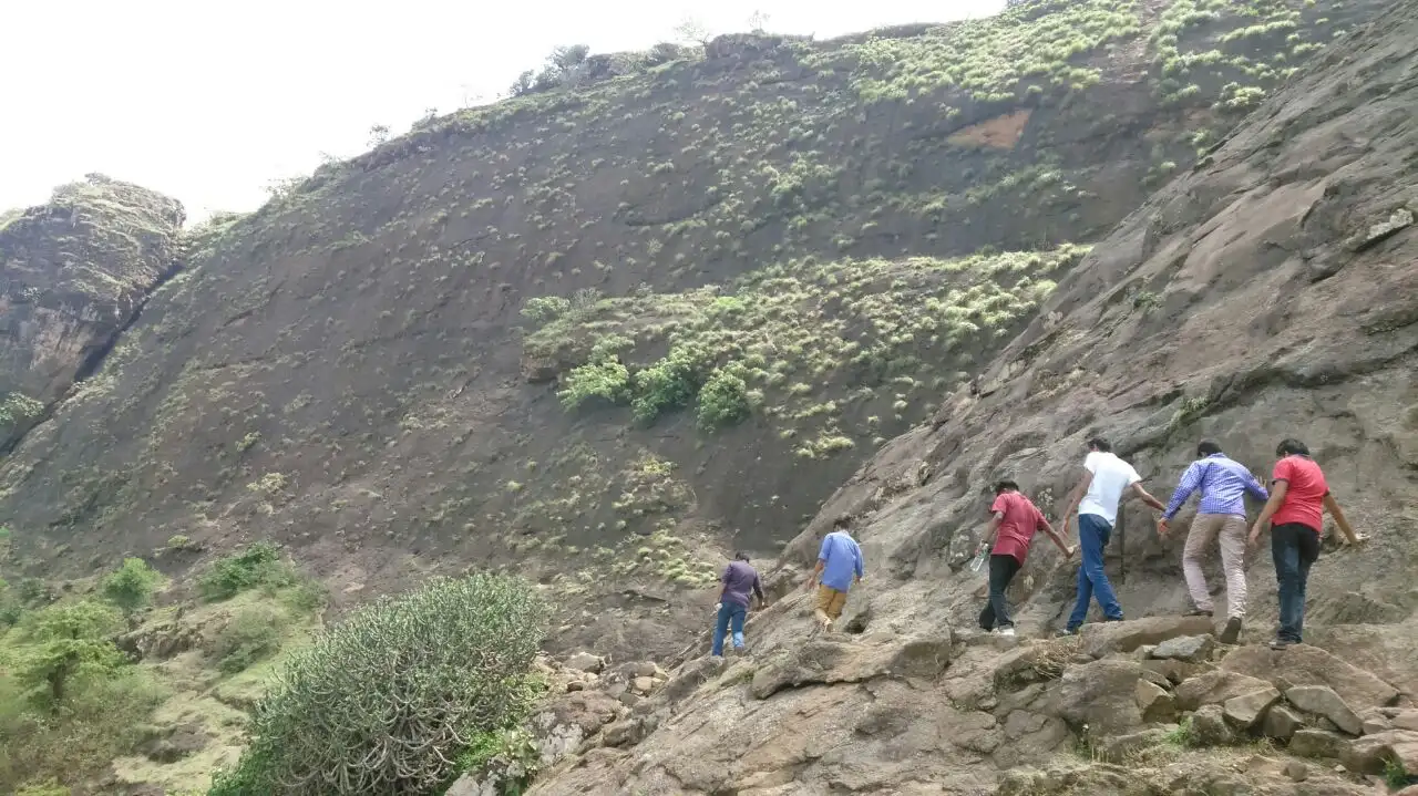 Trekkers climbing a rocky slope below the green-streaked flank of Ramshej hill near Nashik