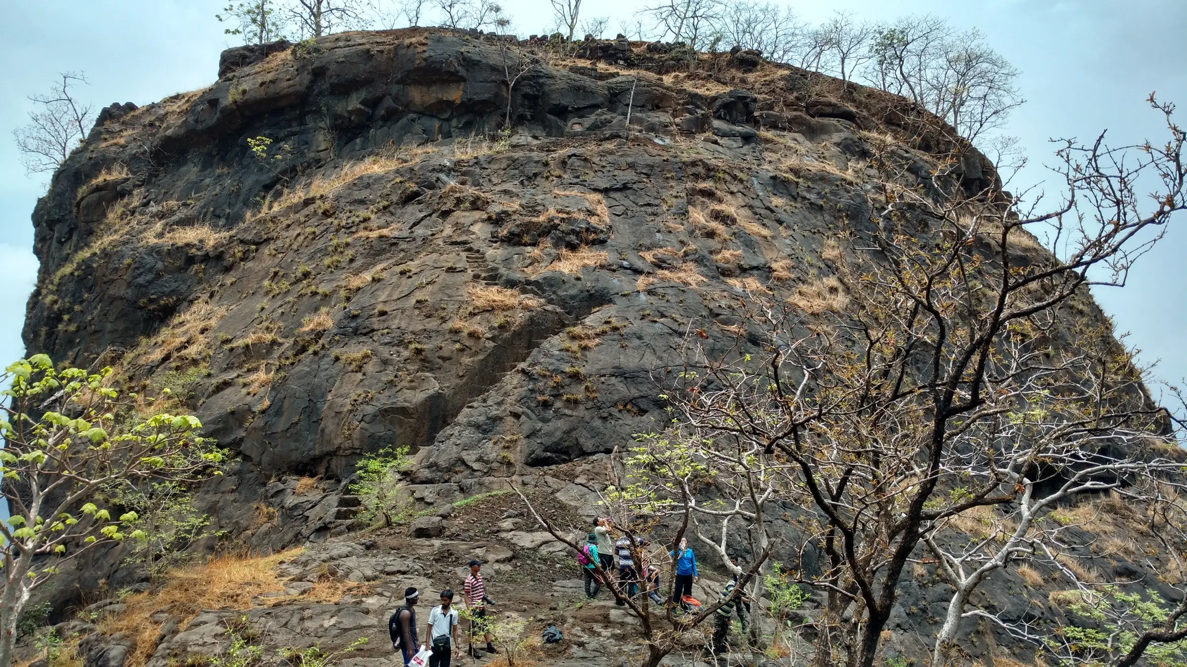Trekkers at the foot of Mrugagad's rock face with rock-cut steps climbing to the top