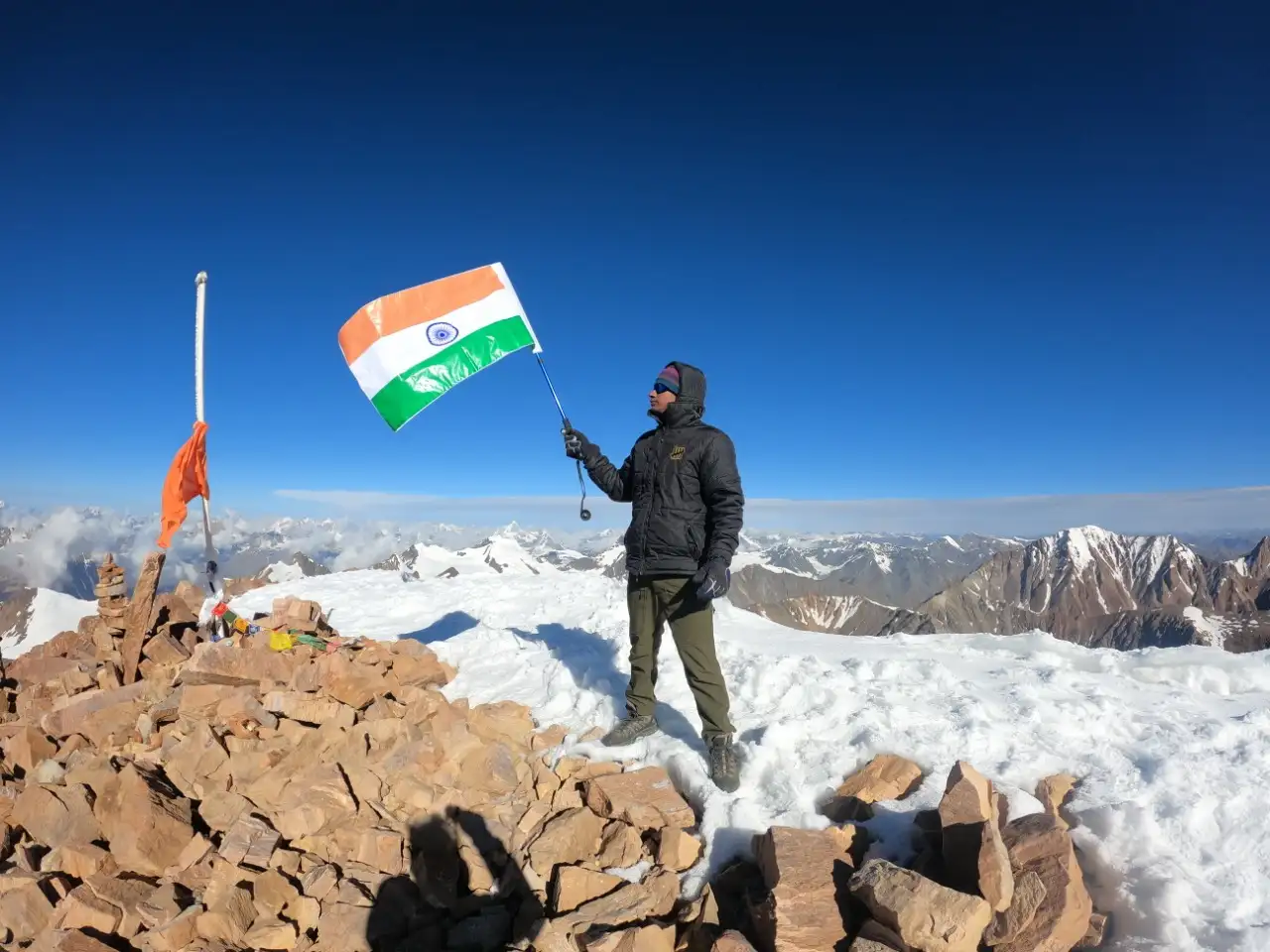 A climber holding the Indian flag beside a rock cairn and prayer flags on a snowy summit the photographer identifies as Mt Yunam, with Lahaul peaks behind