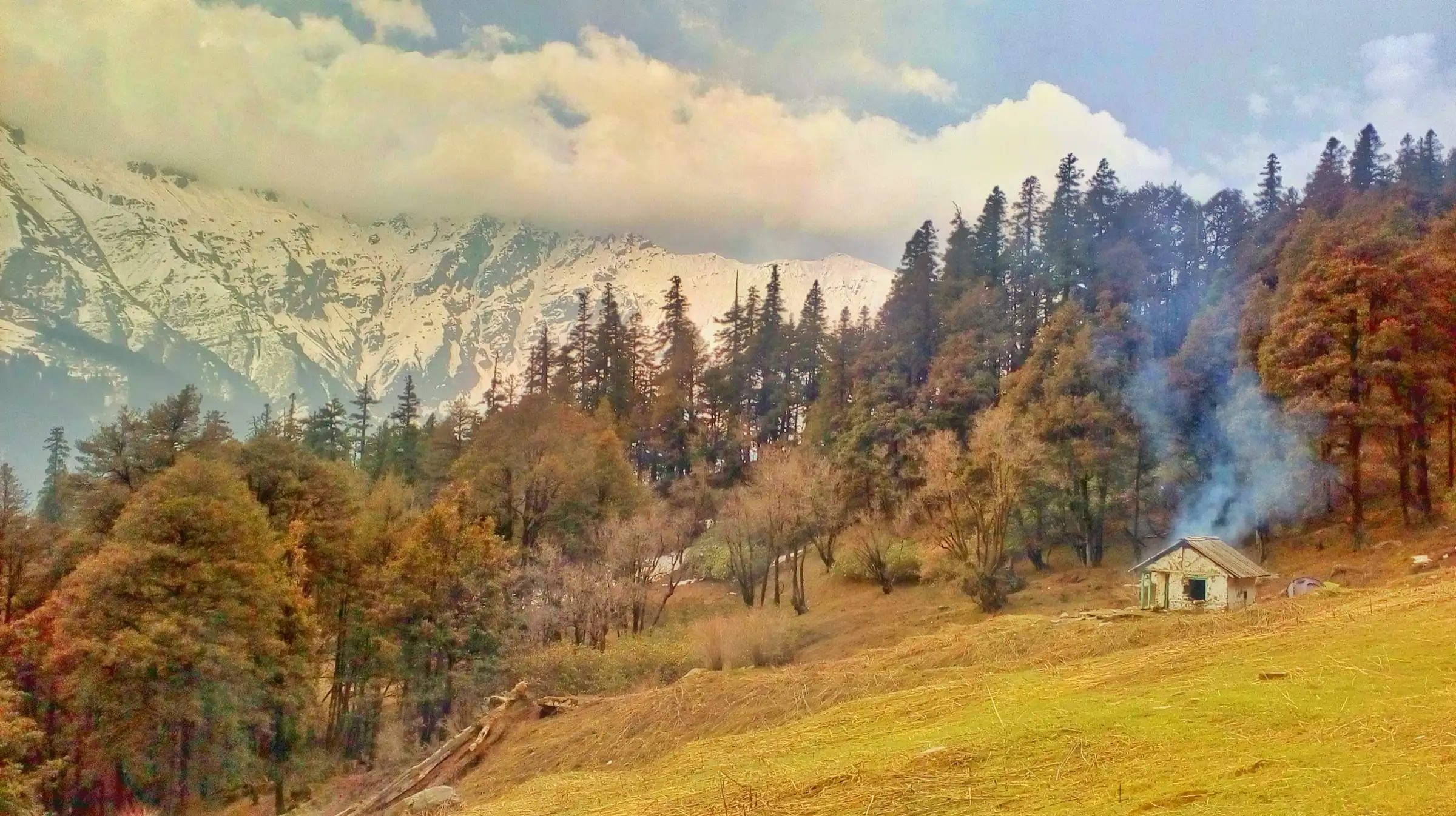 Golden meadow with a small stone hut and smoke rising, conifer forest and snow-streaked ridges under cloud, titled Nada Thach on Commons, a camp on the Tirath route