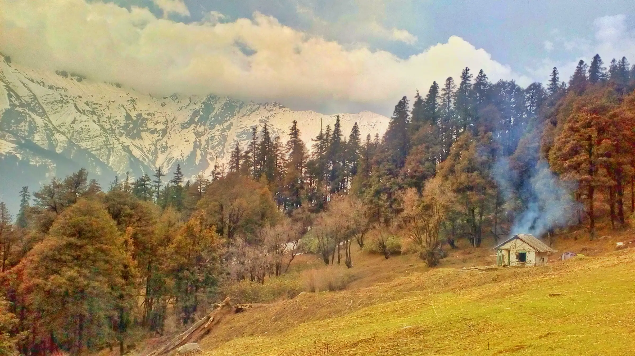 Golden meadow with a small stone hut and smoke rising, conifer forest and snow-streaked ridges under cloud, titled Nada Thach on Commons, a camp on the Tirath route