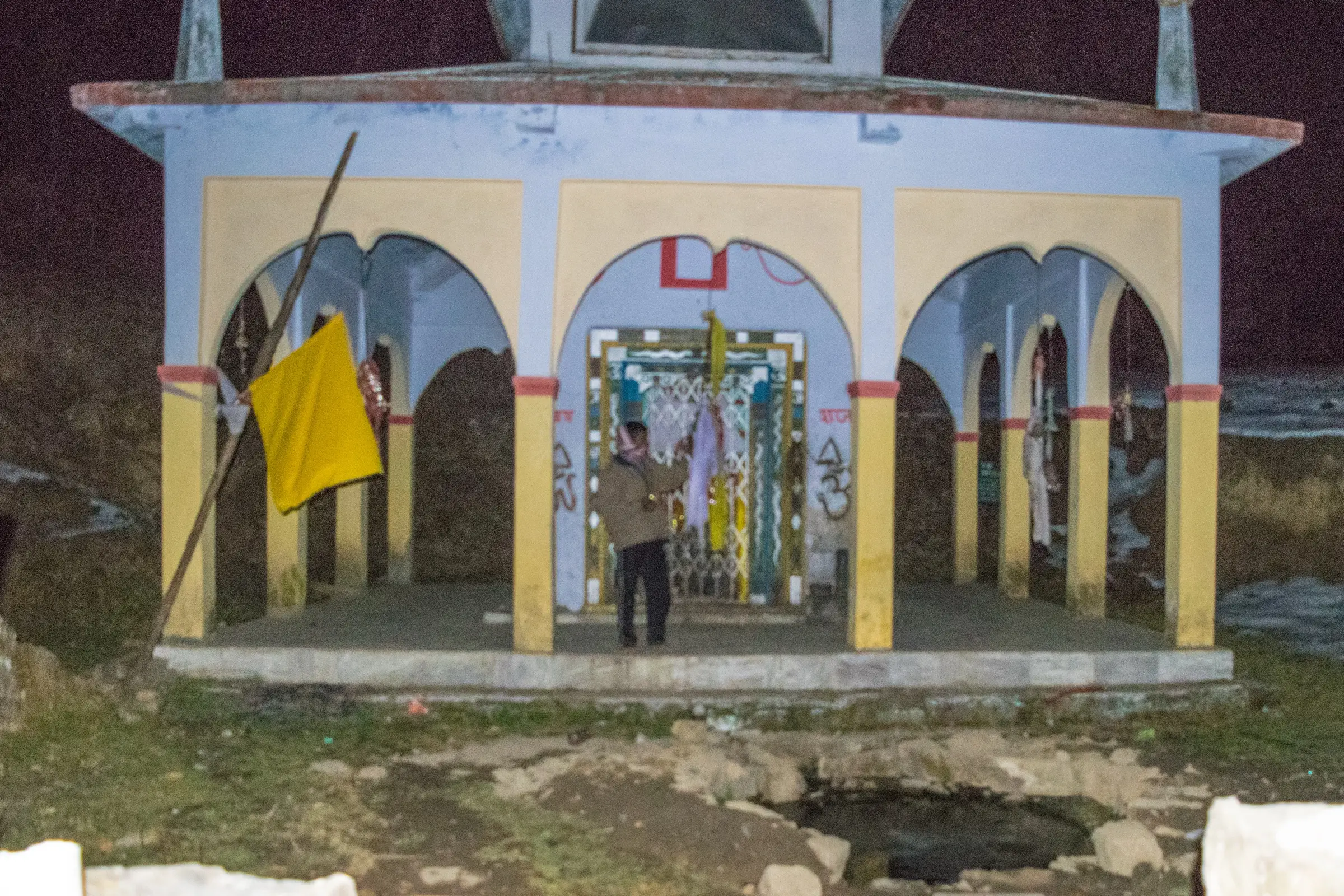 The white and yellow Nag Devta temple with arched pillars and a saffron flag in the forest clearing below Nag Tibba, with its small sacred kund in front